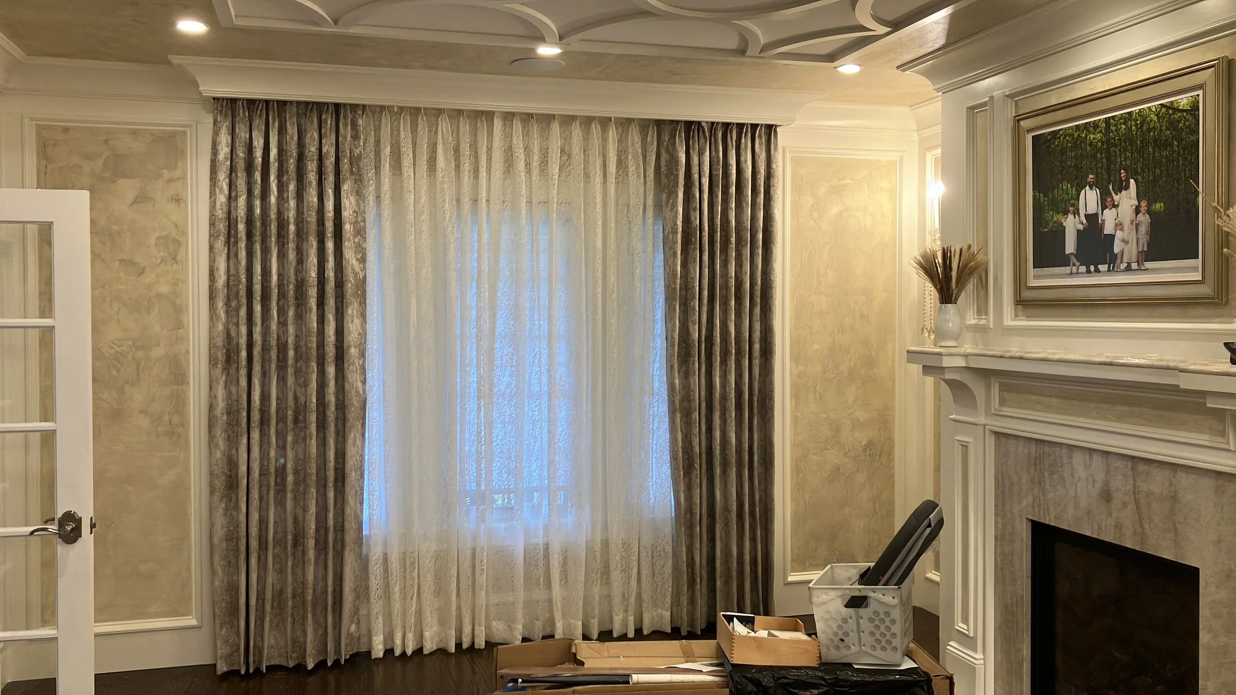 Interior of a living room with beige textured walls, sheer white curtains and heavier patterned curtains on a window, a white mantle fireplace with a picture of a family hanging above, and a black office chair near cardboard boxes.