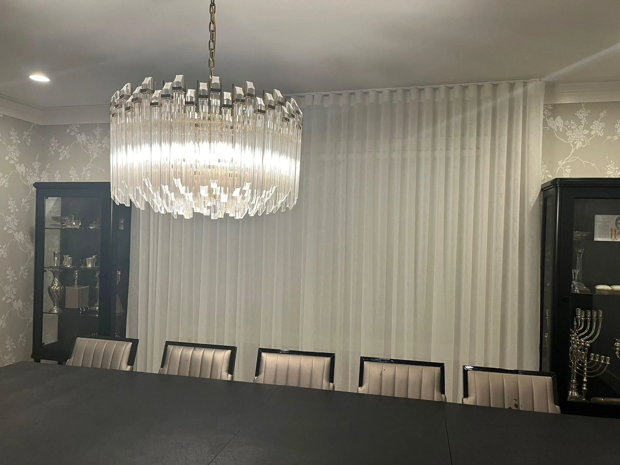 Dining room with a large crystal chandelier, a long table, nine upholstered chairs, and display cabinets with glass doors containing silverware and menorahs.
