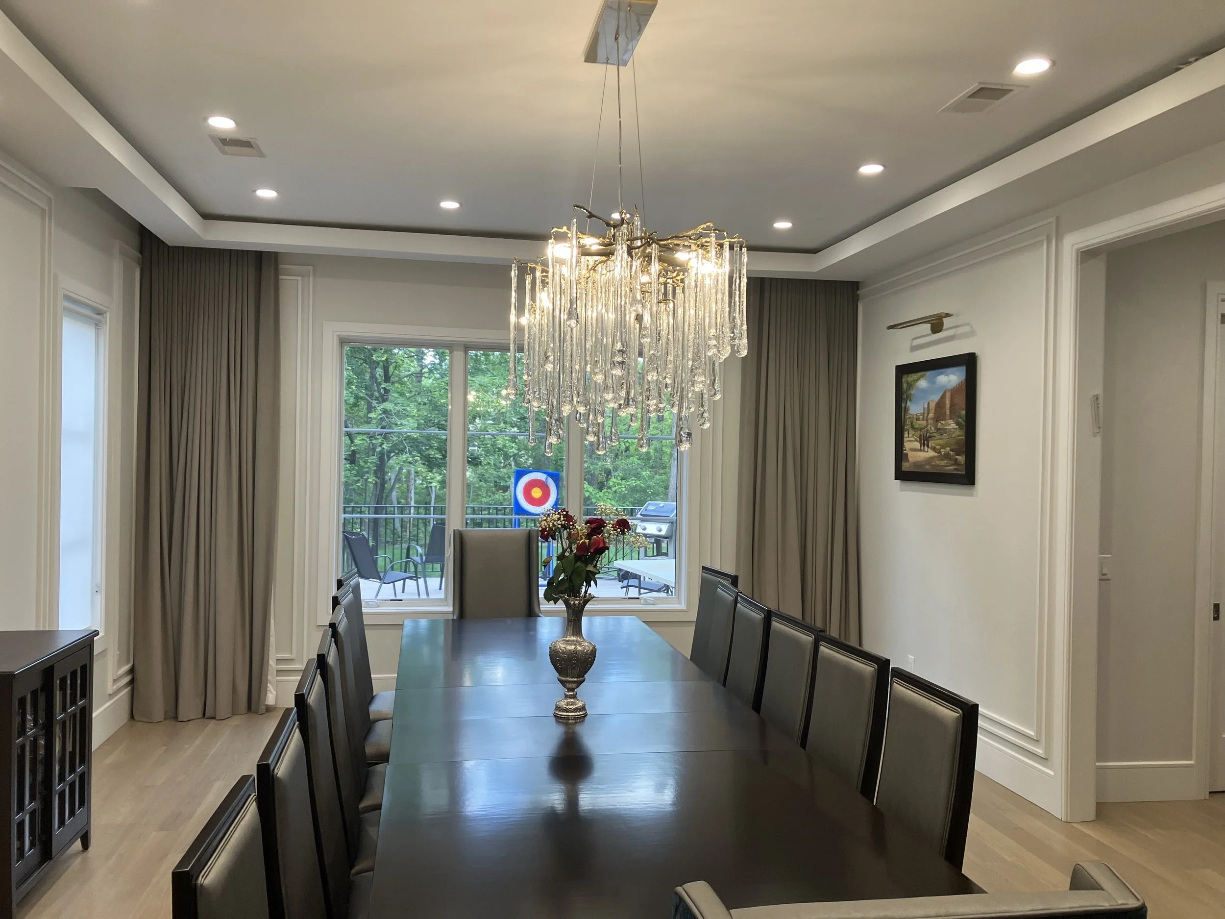 Elegant dining room with a long dark wood table, gray upholstered chairs, a crystal chandelier, and a view of a greenery-filled outdoor patio with a target logo in the background.