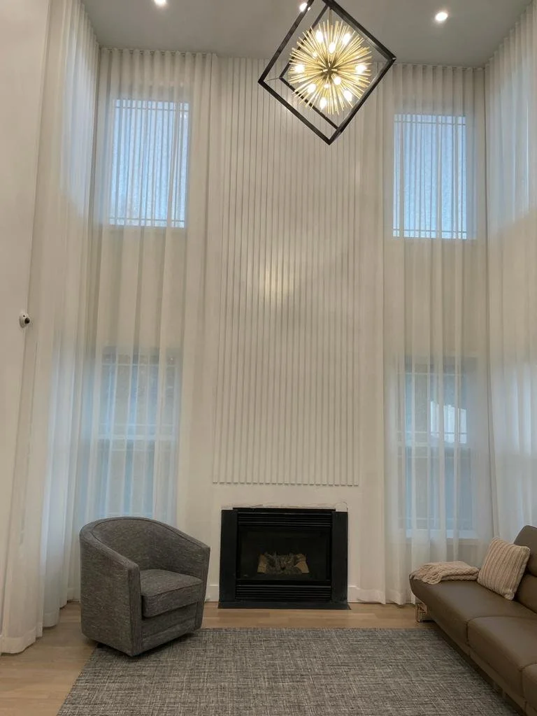 Living room with high white curtains, a modern chandelier, a gray upholstered armchair, a beige sofa, patterned throw pillow, a light-colored area rug, and a black fireplace.