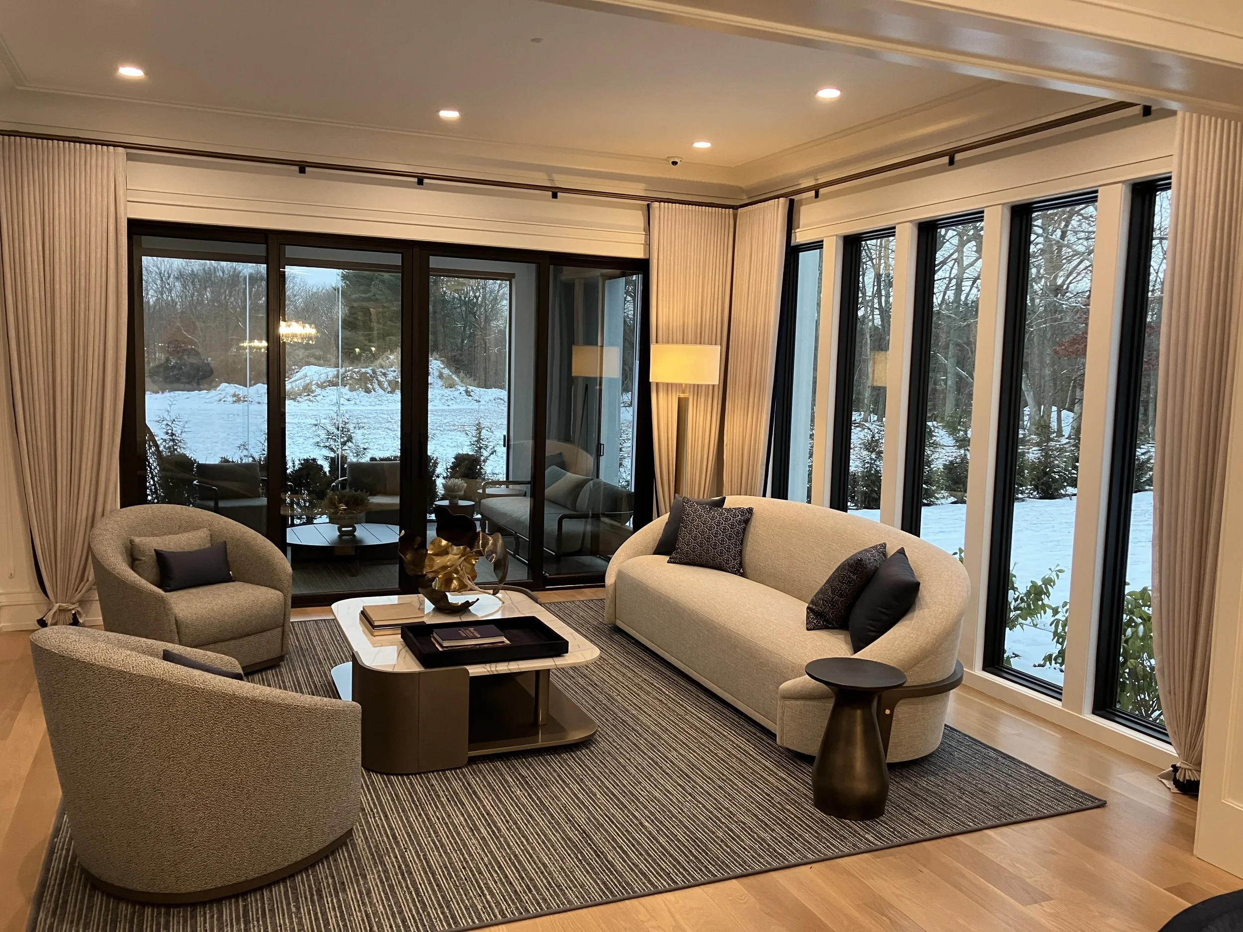 Elegant living room with large windows showing snowy landscape, modern beige sofa with dark pillows, two rounded armchairs, coffee table with decor, and a floor lamp.