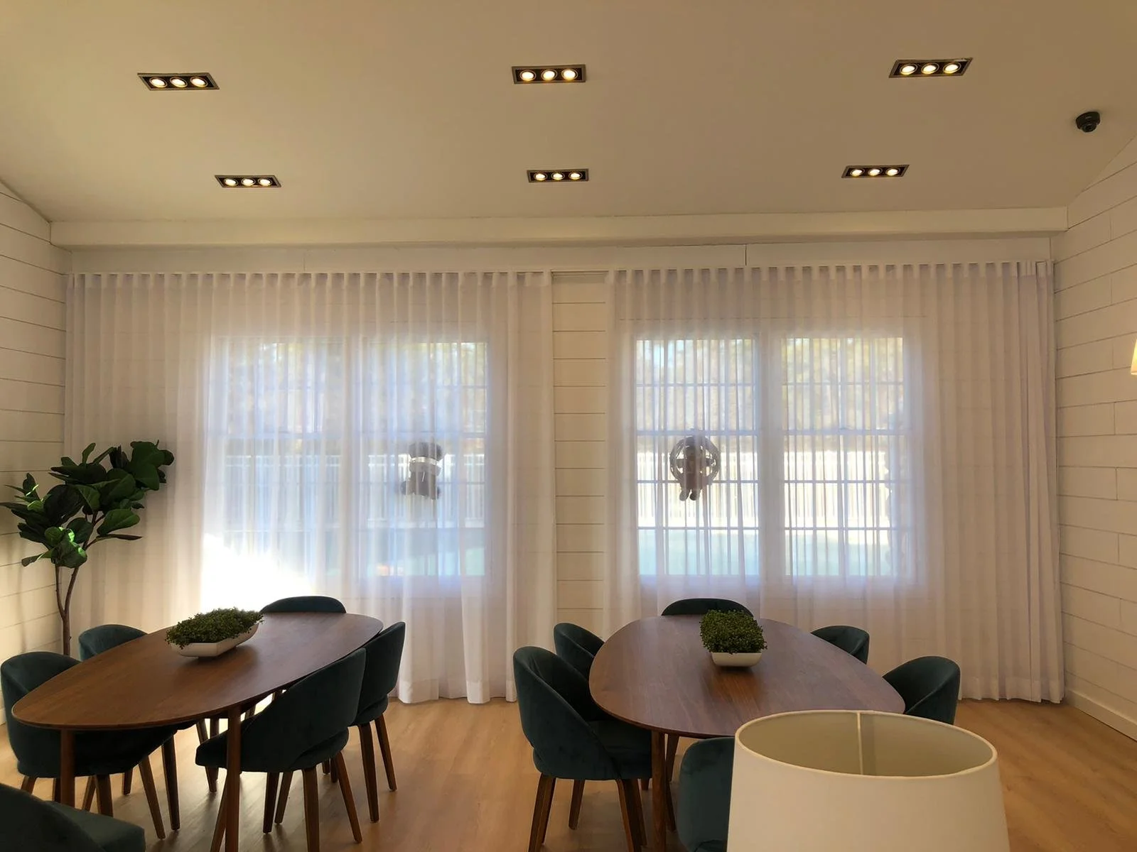 A dining room with two oval wooden tables surrounded by teal upholstered chairs, a potted plant, and a window with sheer white curtains.
