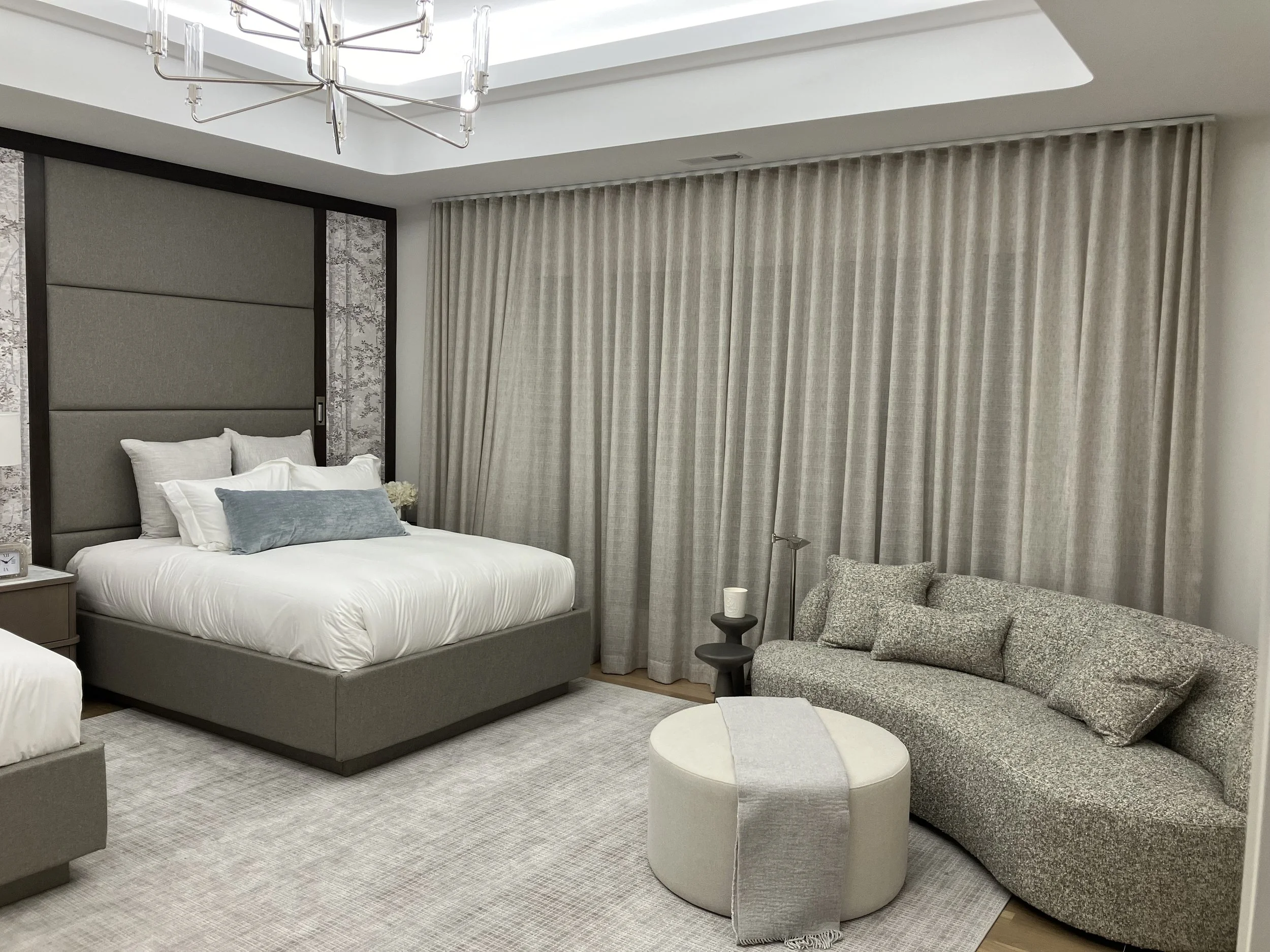 A modern bedroom with a large bed, beige upholstered headboard, gray bedding, white and gray pillows, a small nightstand with a clock, long creamy curtains covering the window, a curved gray sofa with matching pillows, a small round ottoman with a gr