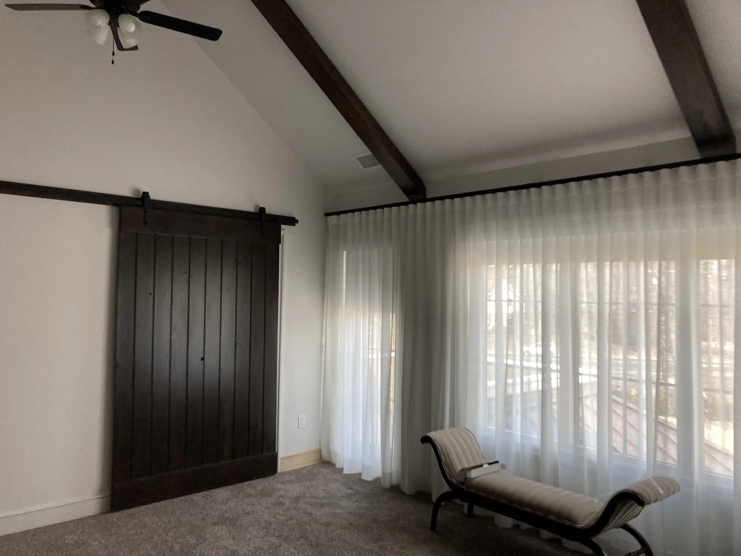 A room with white walls, dark wood beams on the ceiling, a ceiling fan, sheer white curtains on large windows, a wooden sliding door, and a chaise lounge near the window.