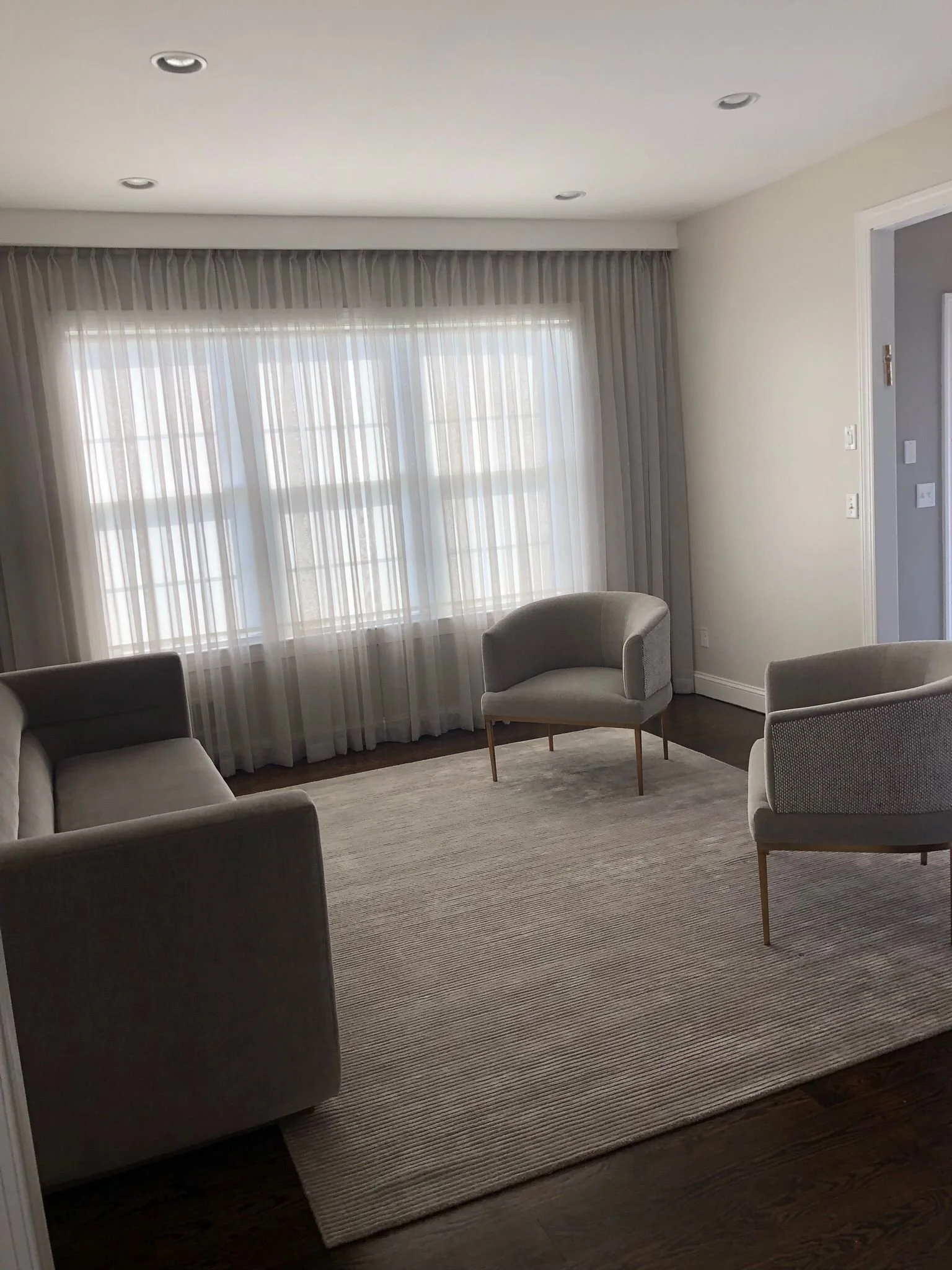 Living room with three modern armchairs and a sofa, a large window with curtains, and a beige area rug on dark hardwood floor.