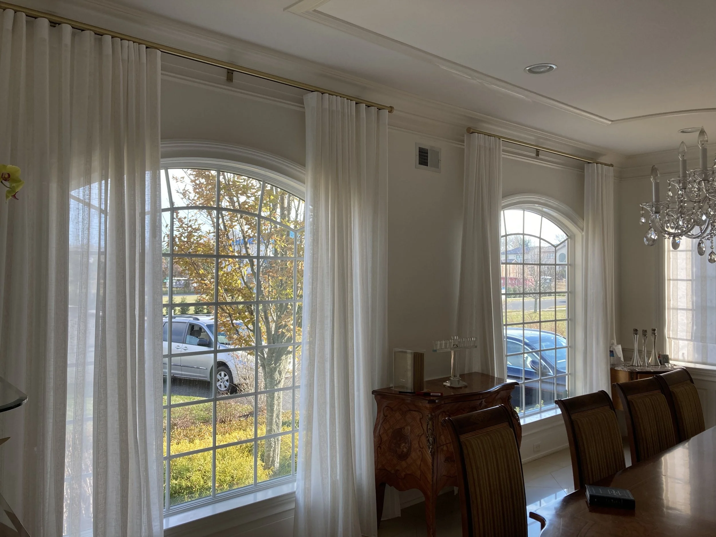Interior of a dining room with large arched windows, white curtains, a wooden dining table with chairs, a chandelier, and a sideboard with decorative items. Outside the windows, trees, a parked car, and a street are visible.