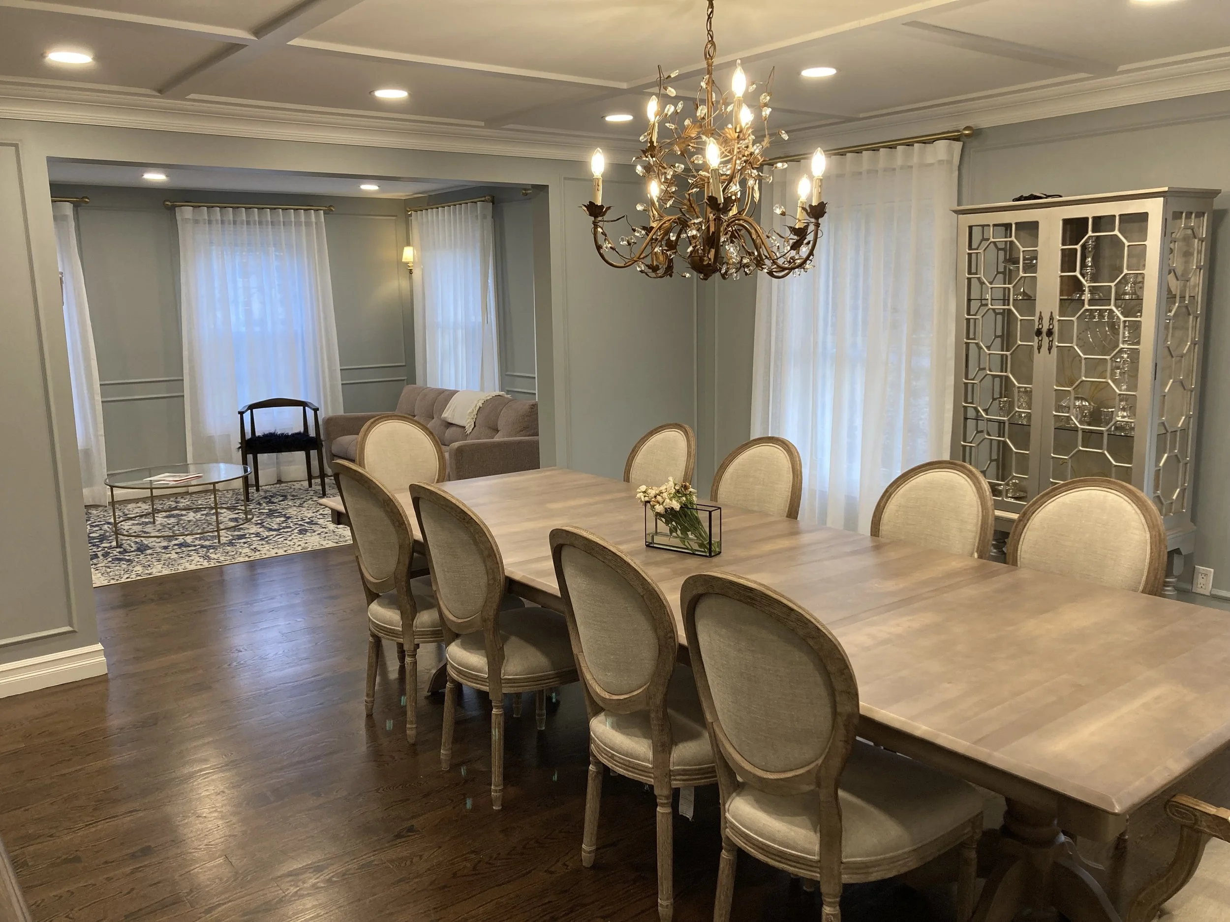 A dining room with a large wooden table surrounded by eight upholstered chairs. A chandelier hangs above the table. There are light-colored curtains on the windows, and a china cabinet in the corner. In the background, a sitting area with a sofa, cha