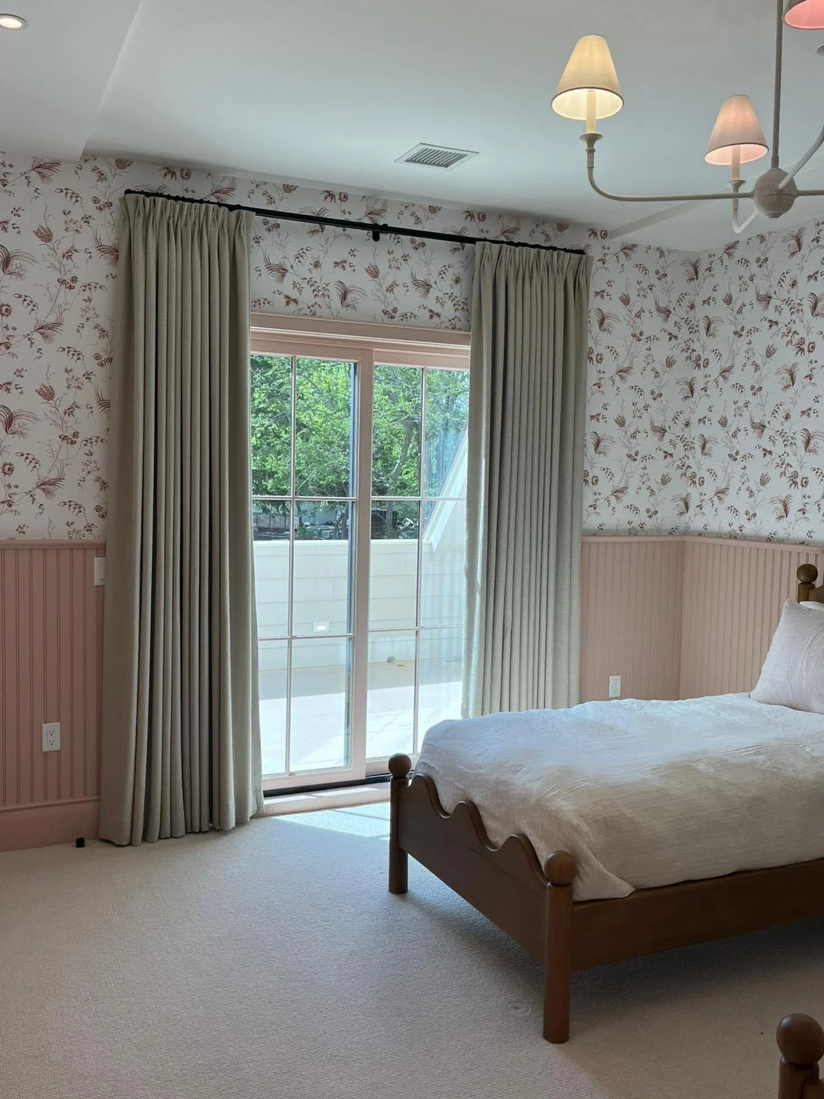 Bedroom with sliding glass door, cream curtains, floral wallpaper, and a wooden bed covered with a white bedspread.