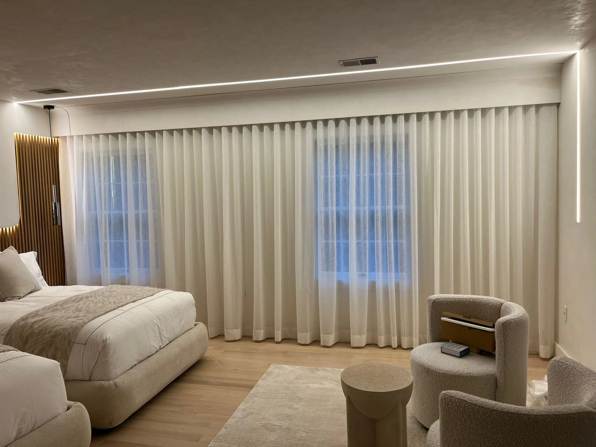 Modern bedroom with beige bedding, sheer white curtains, a cozy armchair, and soft lighting.