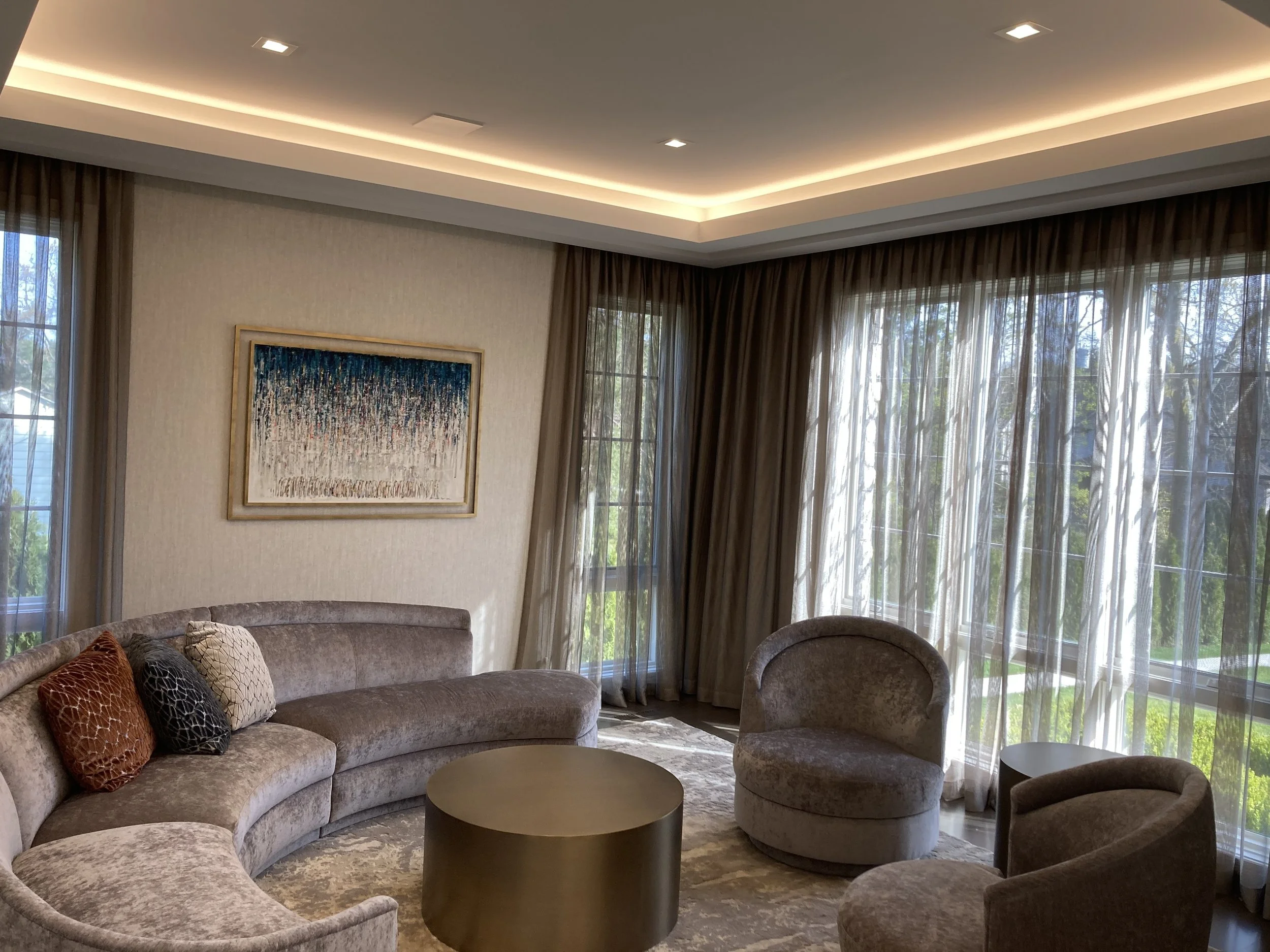 Luxurious living room with a curved sofa, two plush armchairs, a round coffee table, abstract artwork on the wall, and large windows with sheer curtains allowing natural light.