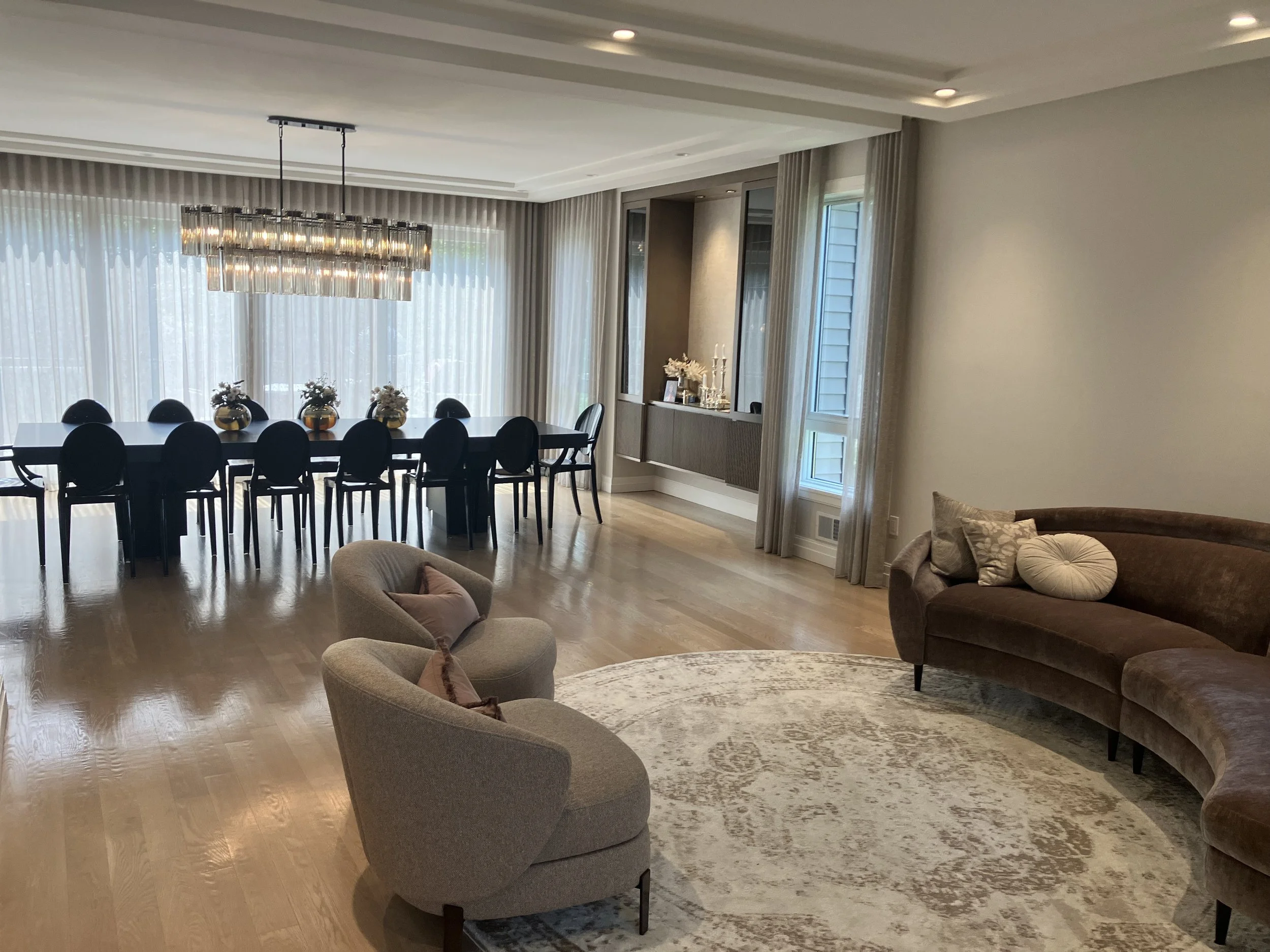 Elegant dining and living room area with a large rectangular dining table surrounded by black chairs, a chandelier overhead, and a curved sofa with pillows, all in a bright space with large windows and light-colored wooden floors.