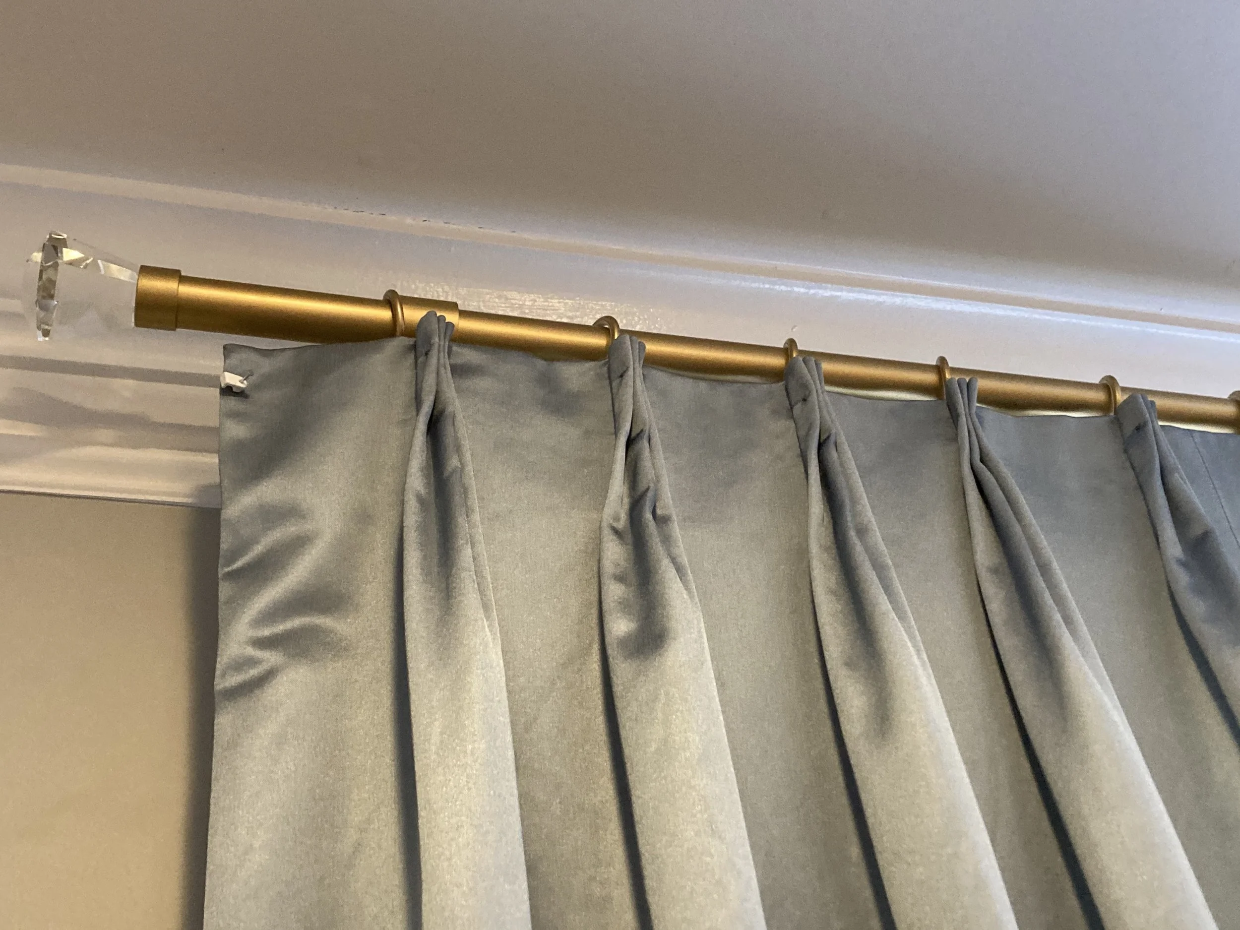 Light gray curtains hanging on a brass curtain rod with a decorative crystal finial, mounted near a white ceiling and beige wall.