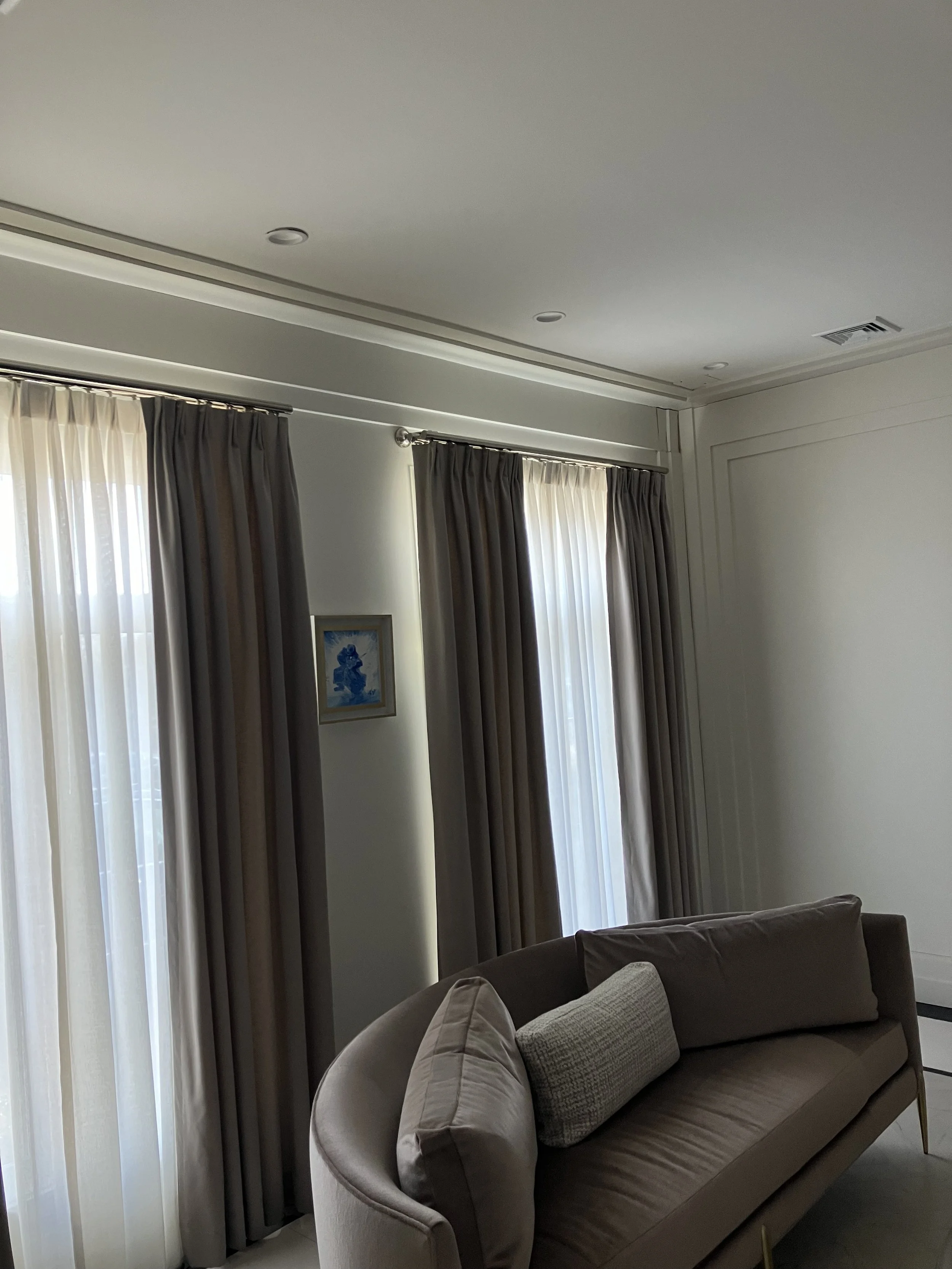 A living room corner with beige curtains, a beige sofa with a cylindrical pillow, and a small framed artwork on the wall.