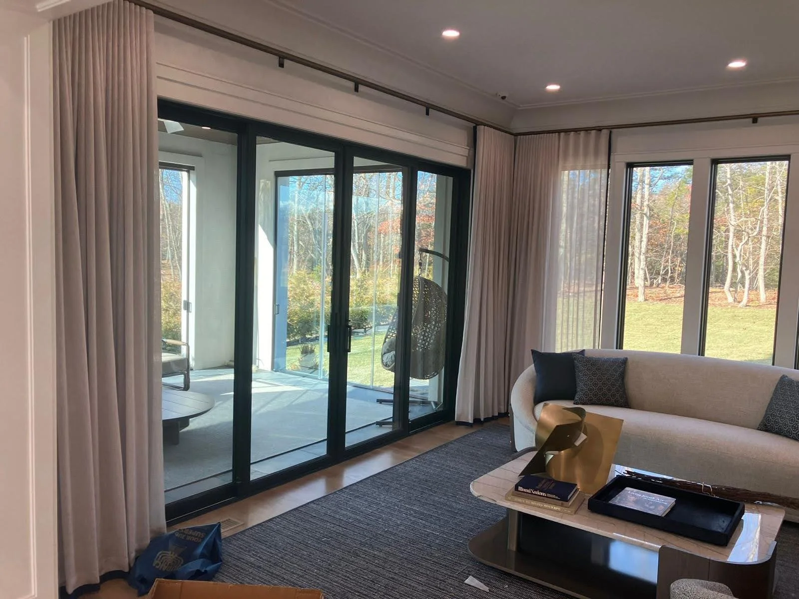 Living room with large glass sliding doors leading to a patio with outdoor furniture, beige curtains, a white curved sofa with throw pillows, a marble coffee table with books and decorative items, and windows showing a backyard with trees.