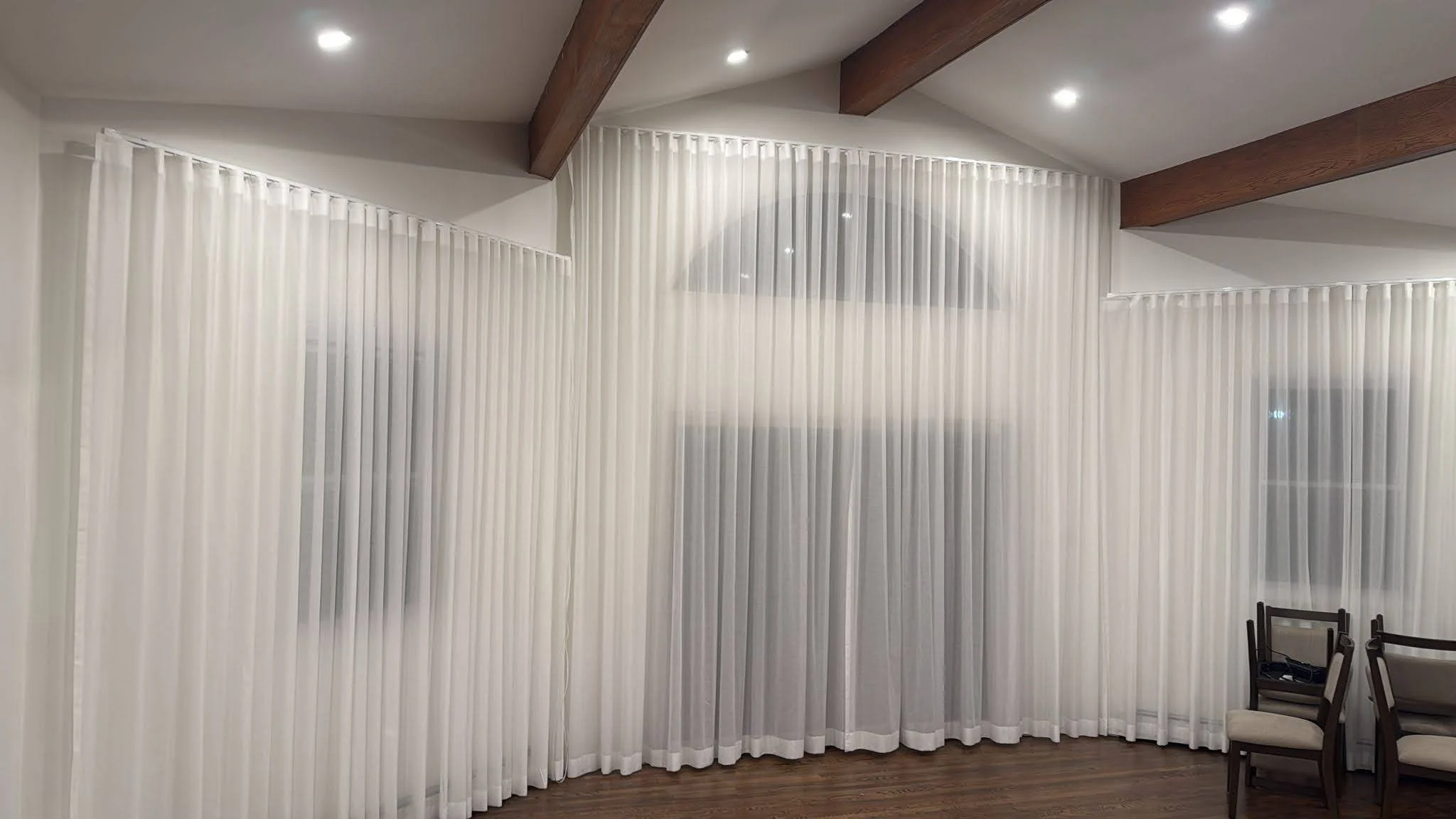 Room with white sheer curtains hanging from ceiling to floor and wooden ceiling beams. There are chairs in the corner of the room.