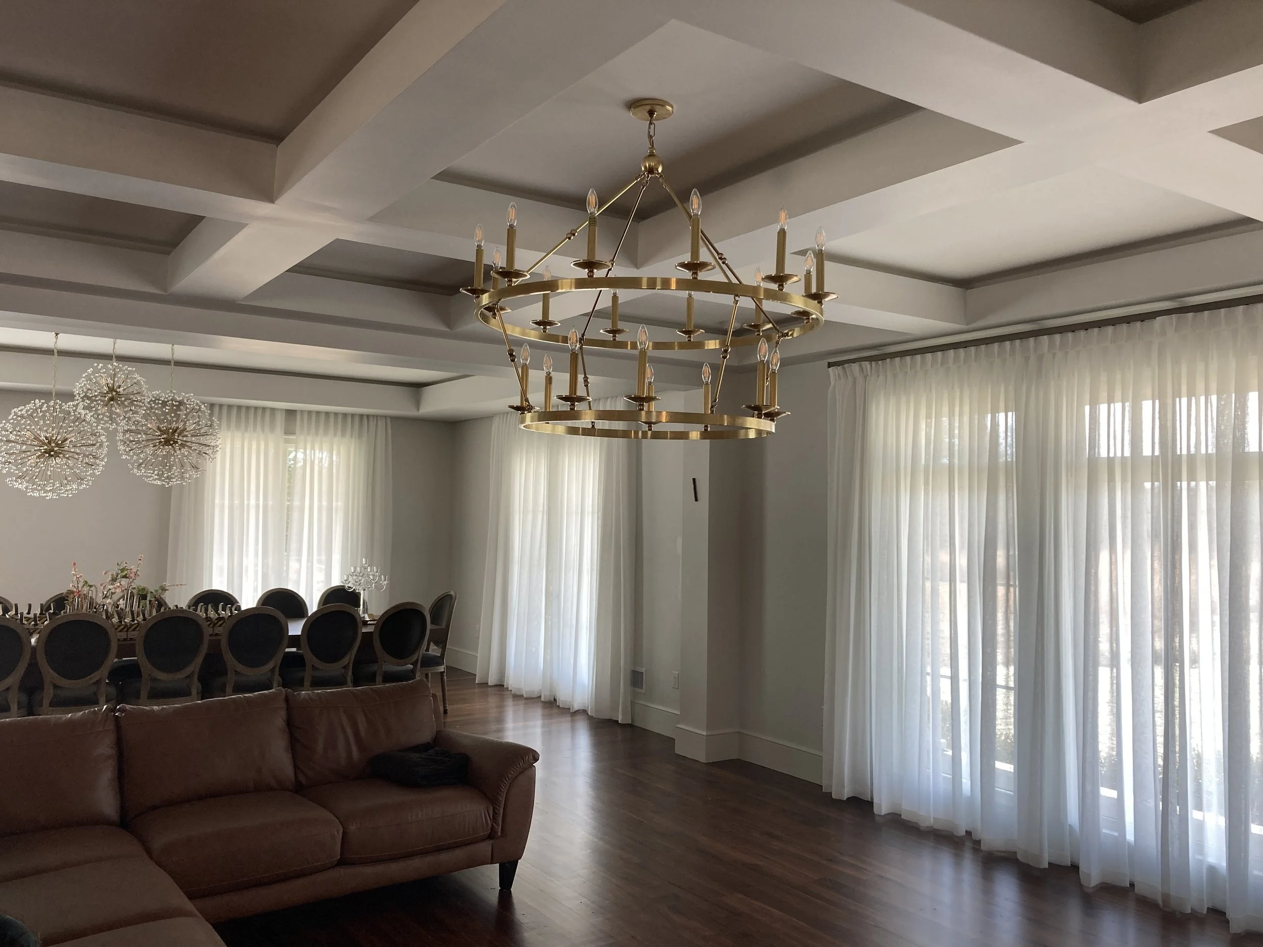 Elegant dining room with a large wooden table, black chairs, floral centerpieces, and modern light fixtures, with large windows covered by white curtains.