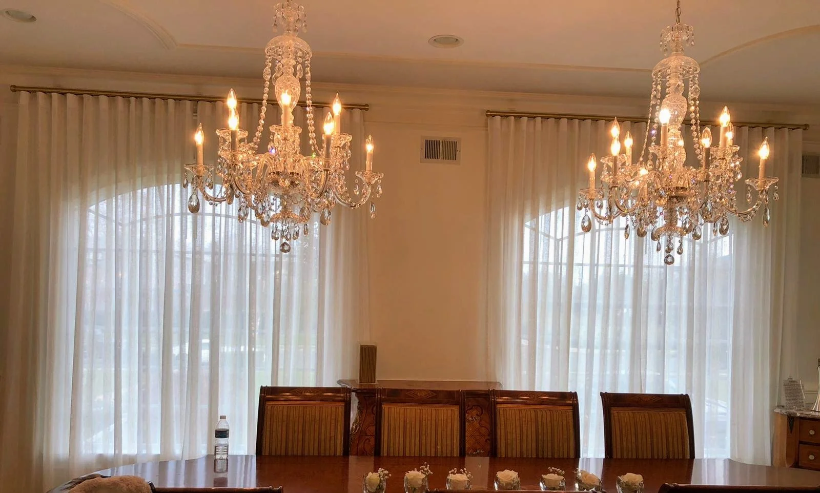 Elegant dining room with two large crystal chandeliers hanging above a wooden dining table, cream-colored curtains behind, and a water bottle and floral table decorations on the table.