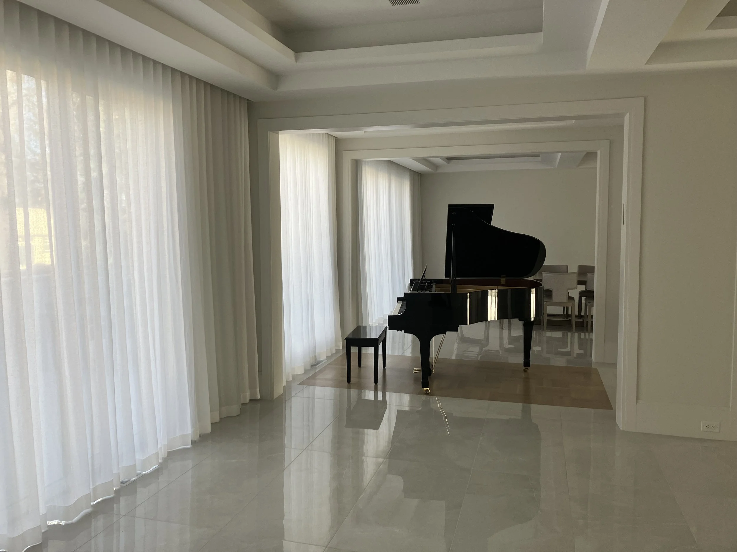 Grand piano in a spacious, well-lit room with white walls, tile flooring, large windows with sheer curtains, and a small sitting area in the background.
