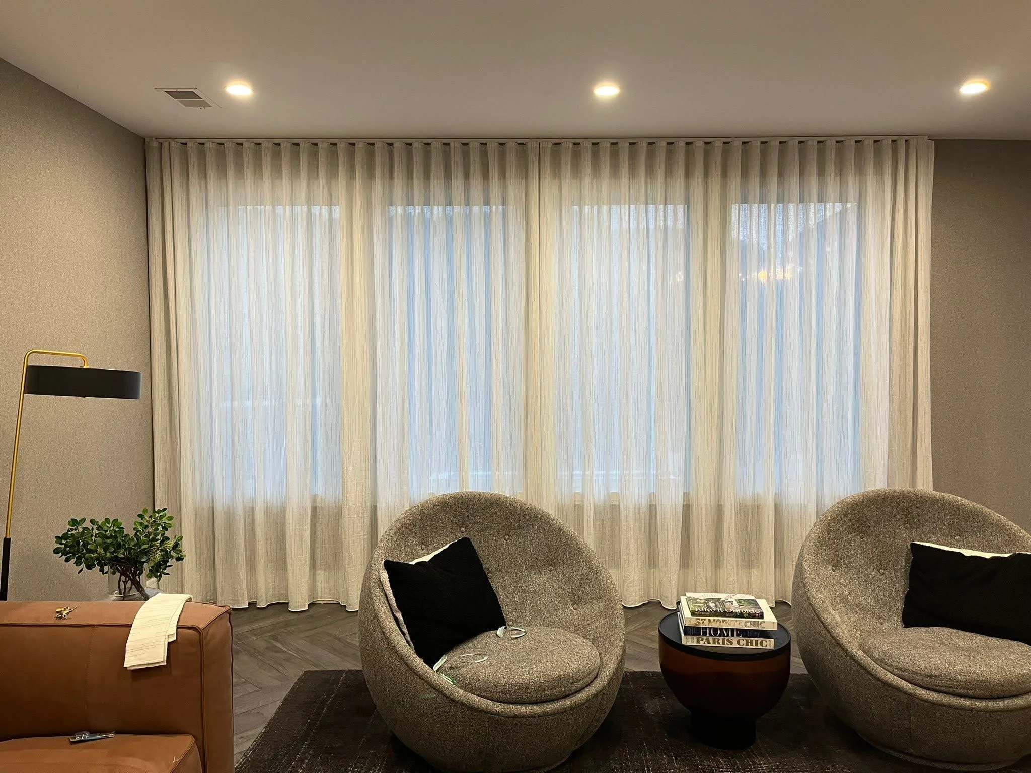 Living room with beige armchairs, black pillows, side table with books, and sheer curtains over large windows