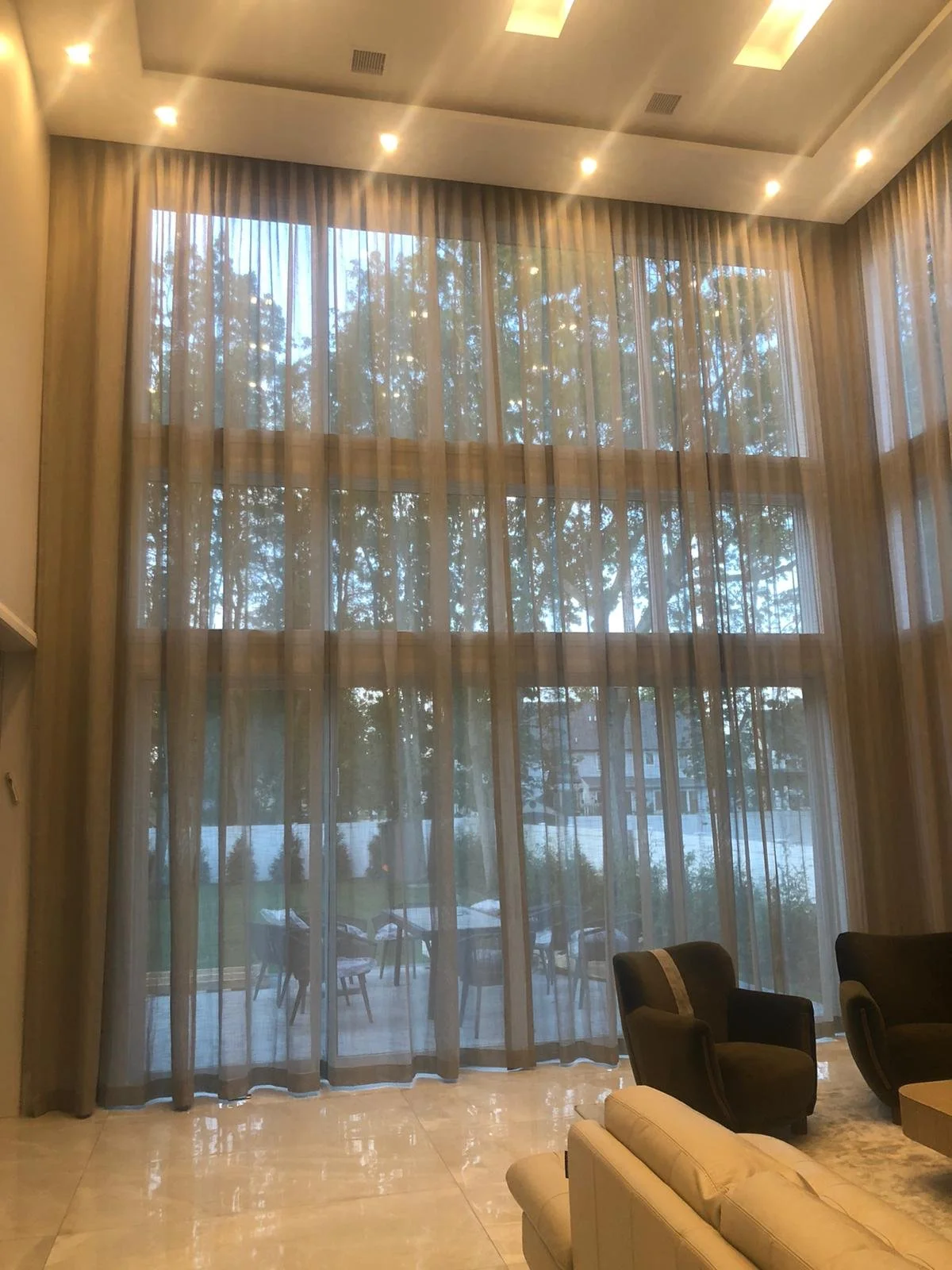 Large window with sheer curtains looking outside to trees and a patio with chairs and a lake in the background. Interior includes beige tiled floor and dark armchairs.