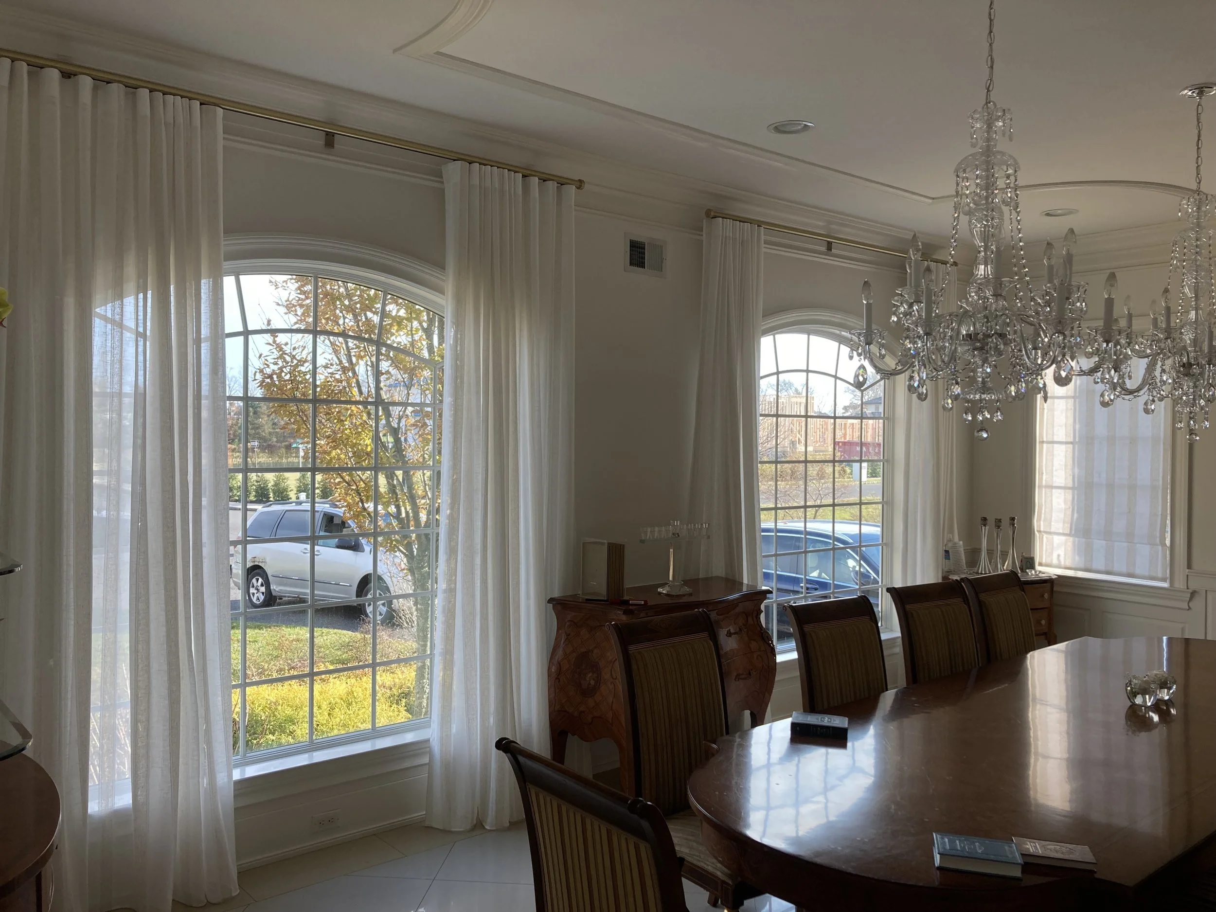 Elegant dining room with large arched windows, white curtains, a wooden oval table, and a crystal chandelier.