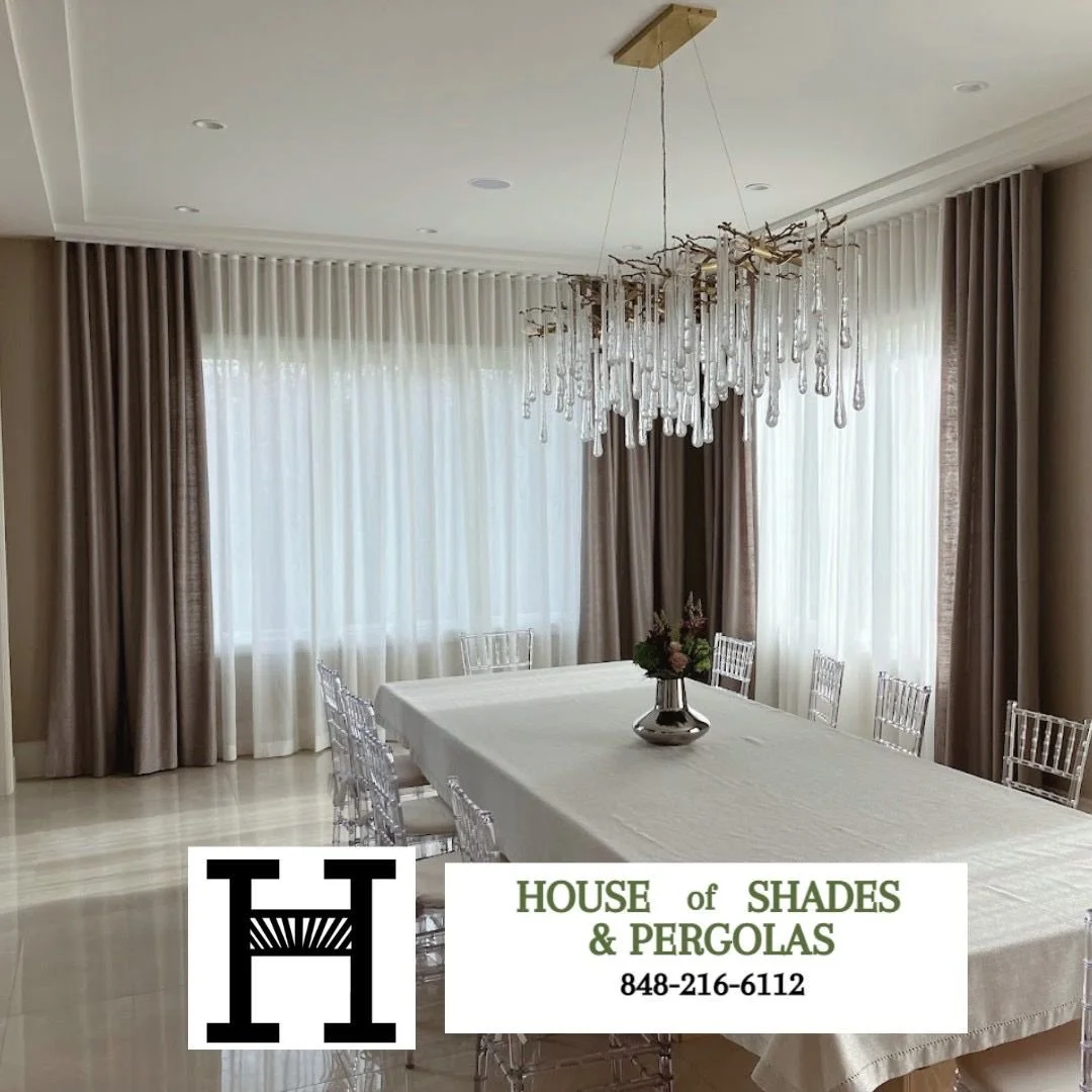 A dining room with a large rectangular table covered with a white tablecloth, surrounded by transparent chairs. A modern chandelier with hanging glass droplets is centered above the table. The room features large windows with sheer white curtains and