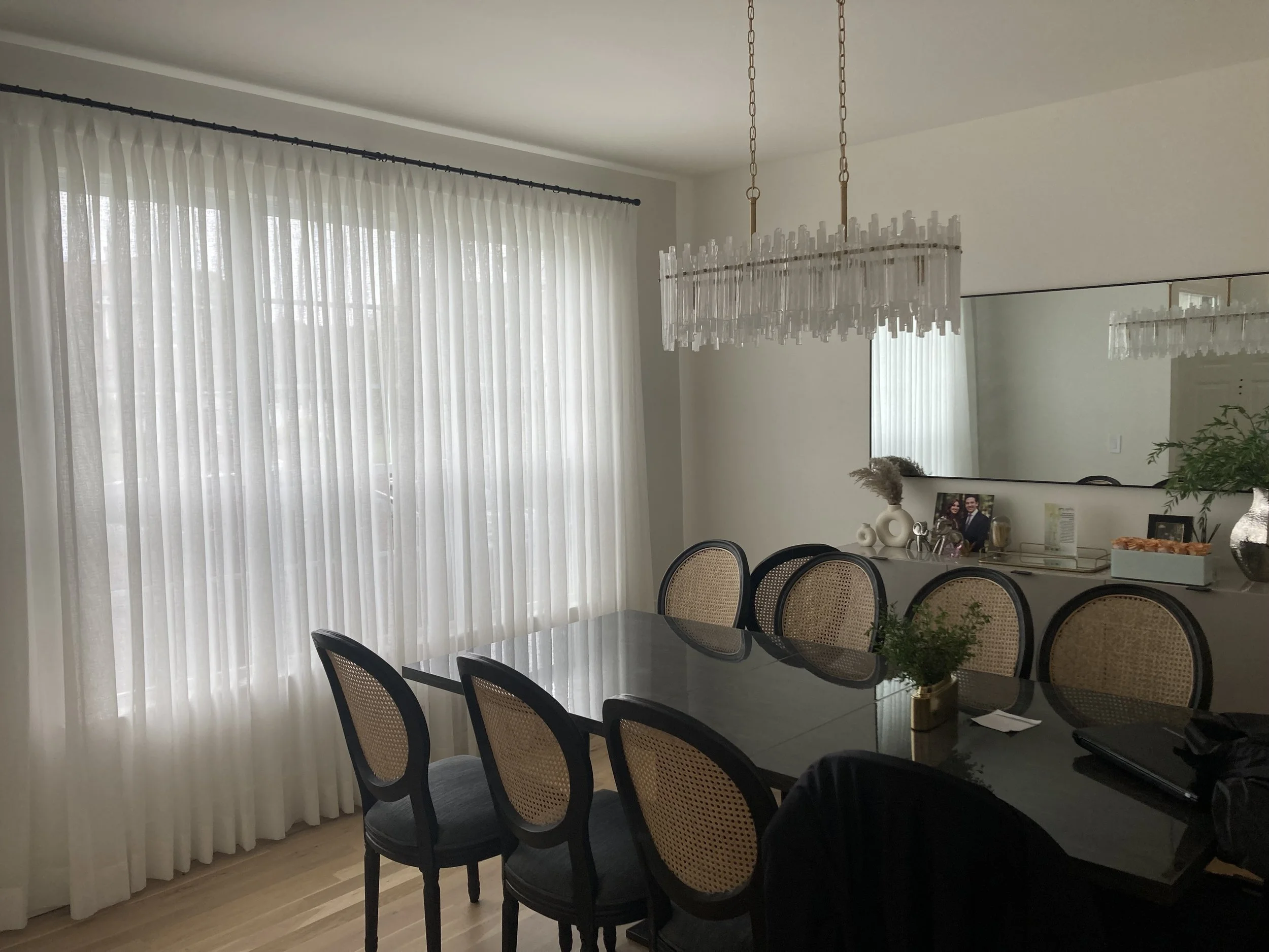 Dining room with black dining table, six cane-backed chairs, large window with sheer white curtains, modern chandelier, and a sideboard with decorative items and a large mirror.