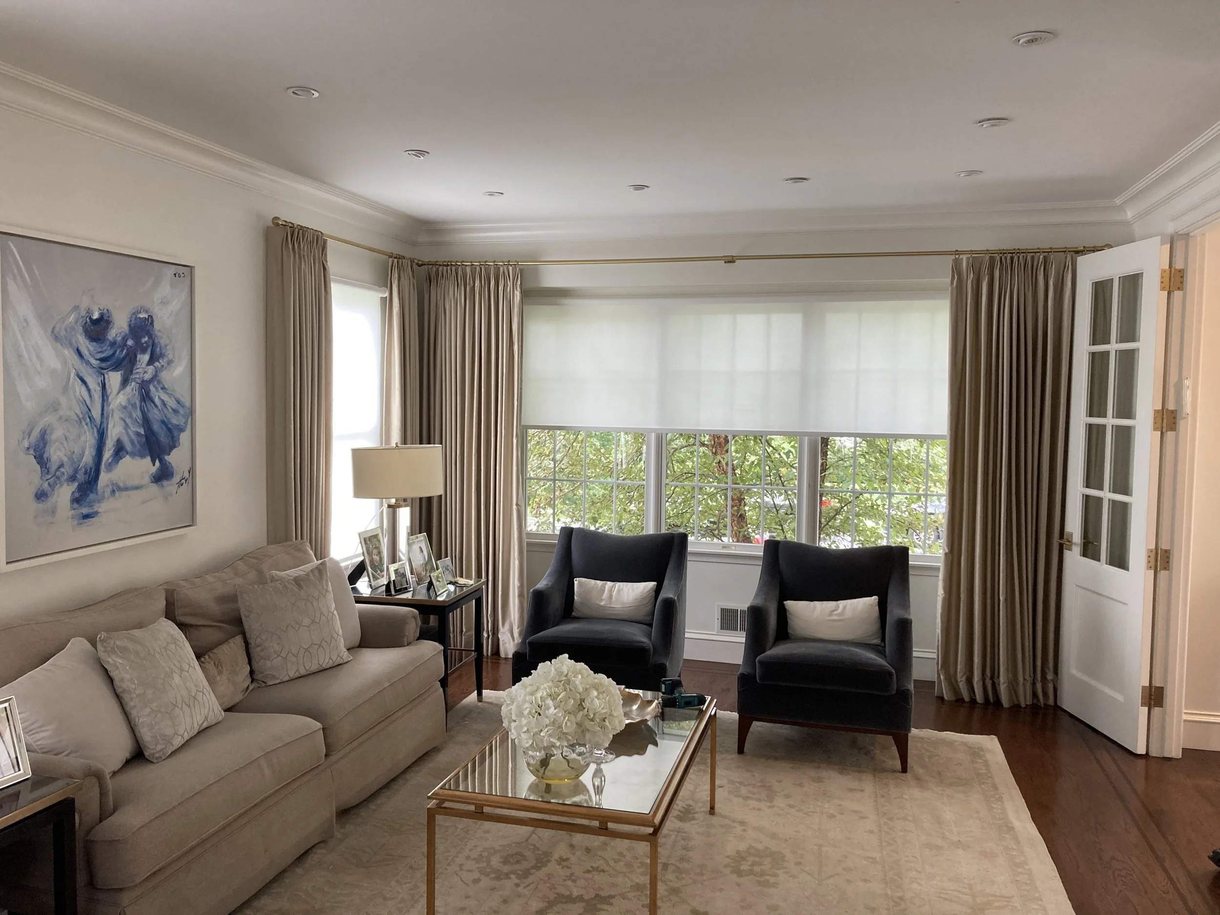 Living room with beige sofa, two black armchairs, a glass coffee table with a vase of white flowers, framed photographs, and a large window with cream-colored curtains and blinds.