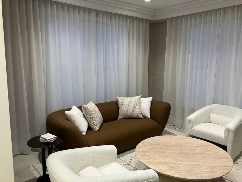 Living room with a brown sofa, white armchair, small black side table with books, round wooden coffee table, and sheer white curtains.