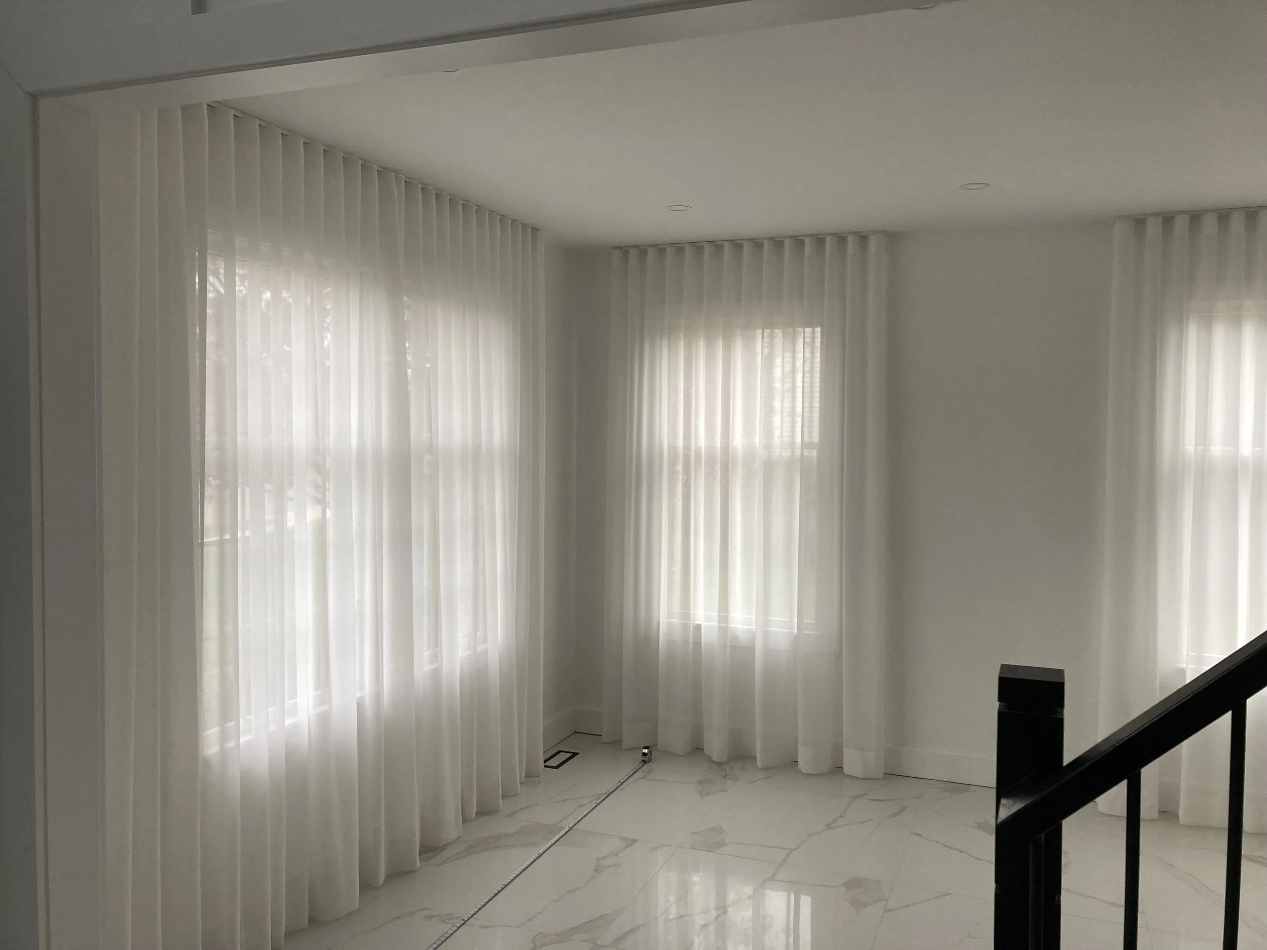 Interior of a bright room with white curtains, marble flooring, and a staircase railing in the corner.