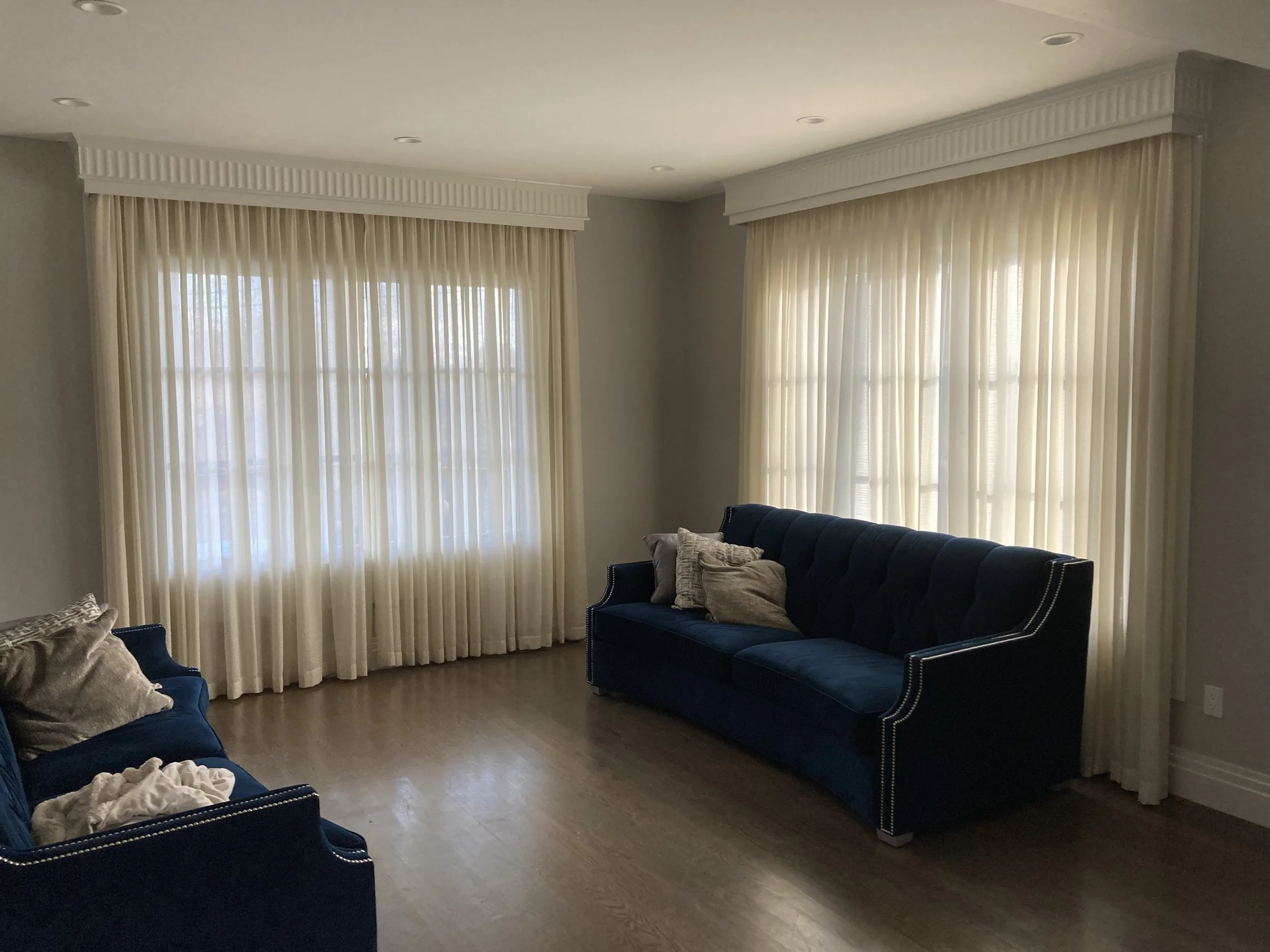 Living room with two large windows covered by cream-colored curtains, a navy blue velvet sofa with multiple throw pillows, and hardwood floors.