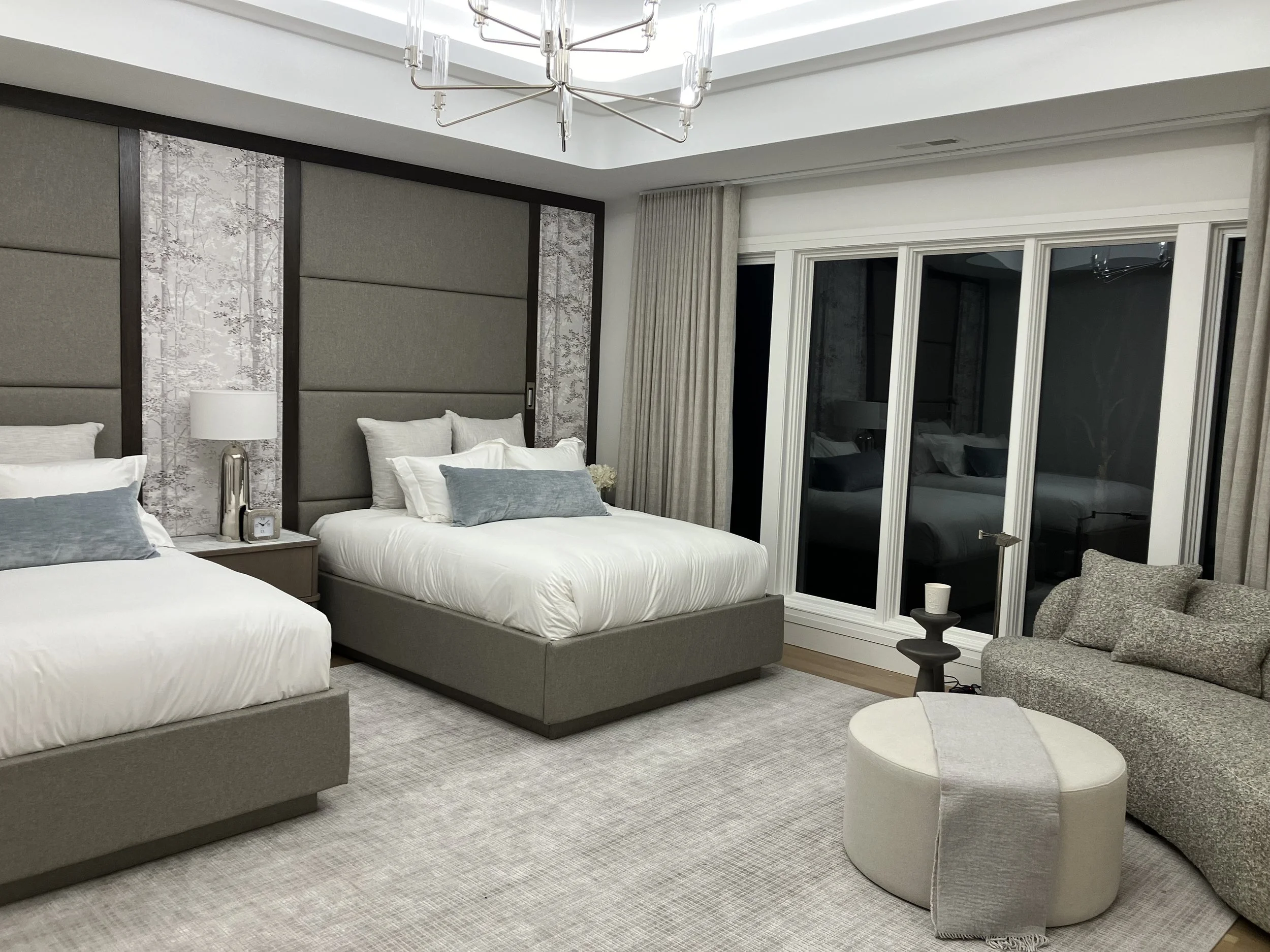 A modern hotel bedroom with two beds, a gray upholstered headboard, a nightstand with a lamp and clock, a sitting area with a gray sofa, a small black side table, an oval ottoman with a blanket, and large windows with beige curtains.