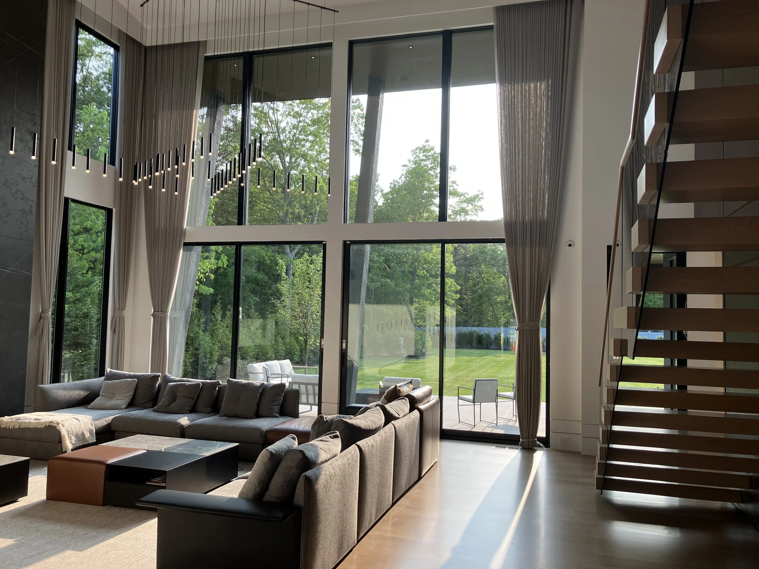 Modern living room with large floor-to-ceiling windows, gray sectional sofa with pillows, black and brown coffee tables, and staircase with wooden steps on the right. Outside, there is patio furniture and a green lawn with trees.