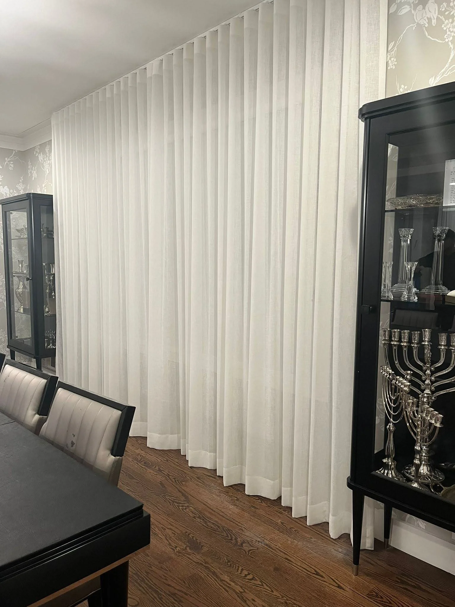 White curtains hanging in a room with dark wood flooring, black display cabinets with glass containing silver and crystal items, and a black dining table with chairs.
