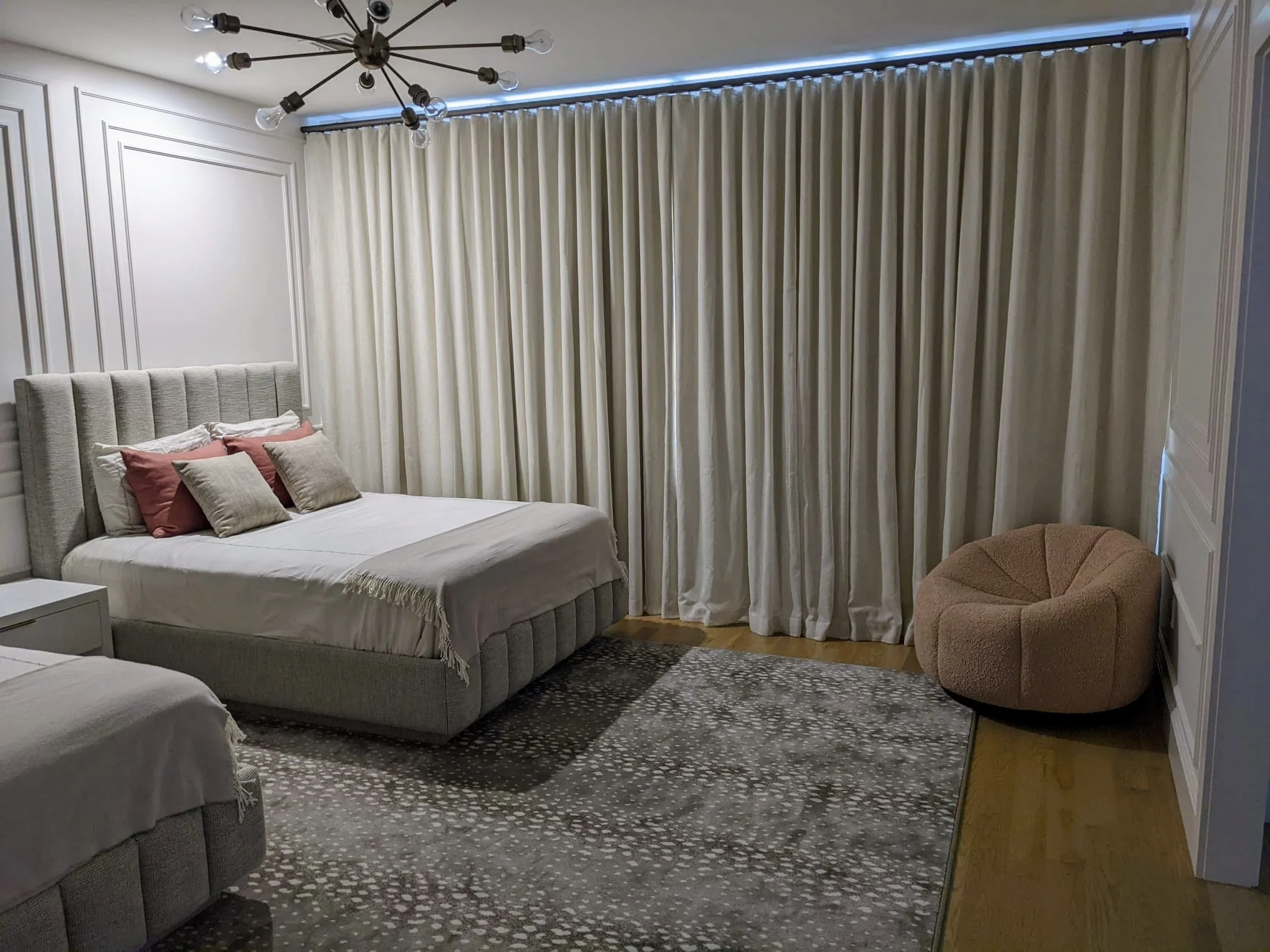 A bedroom with beige curtains covering a large window, a bed with a gray upholstered headboard, white bedding, and decorative pillows, a modern black chandelier with exposed bulbs, a patterned area rug, and a beige round armchair in the corner.