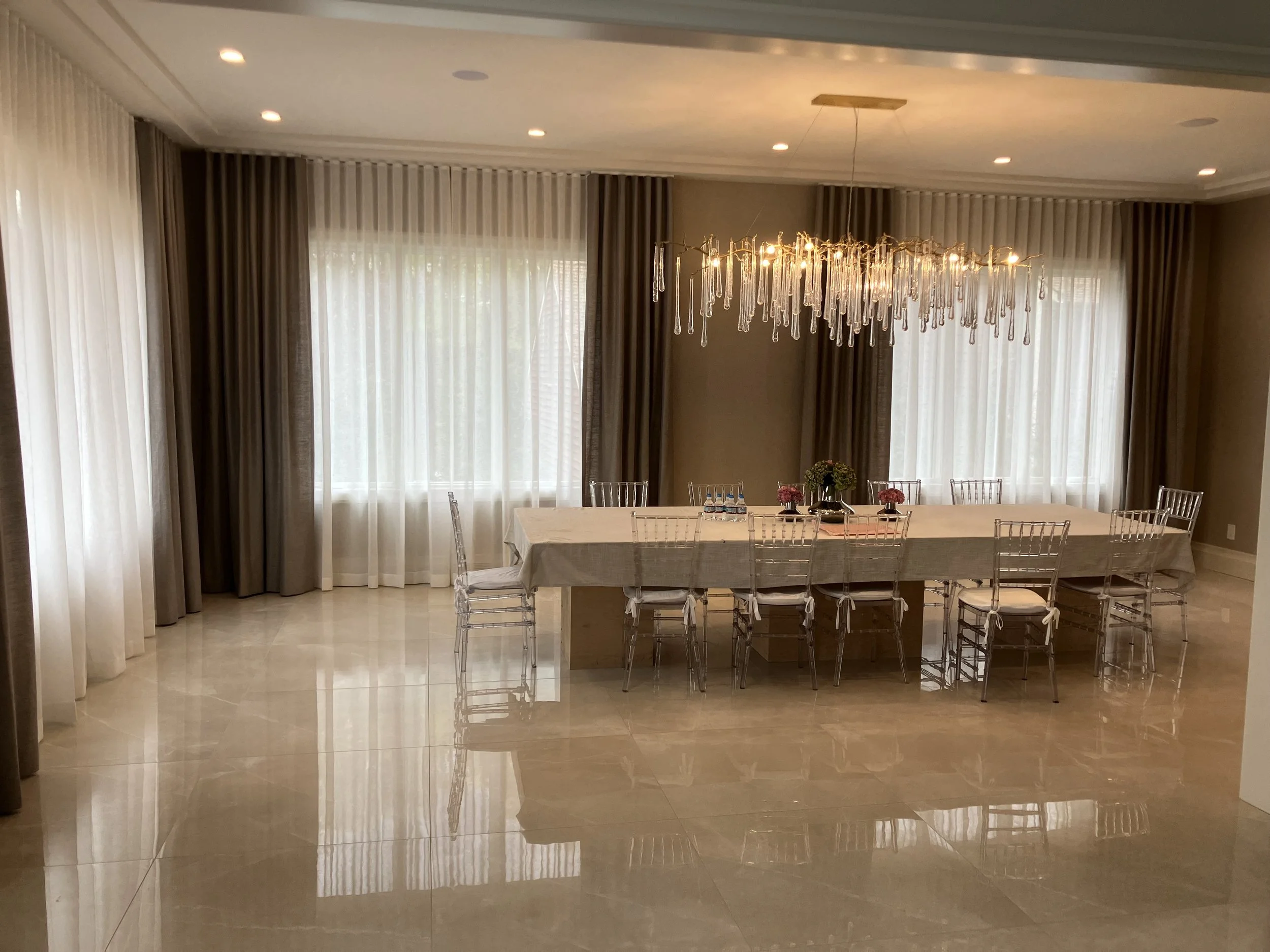 Elegant dining room with a long table covered in a white tablecloth, surrounded by transparent chairs with white cushions. A chandelier with hanging glass elements is above the table, and floor-to-ceiling curtains are in the background, with natural