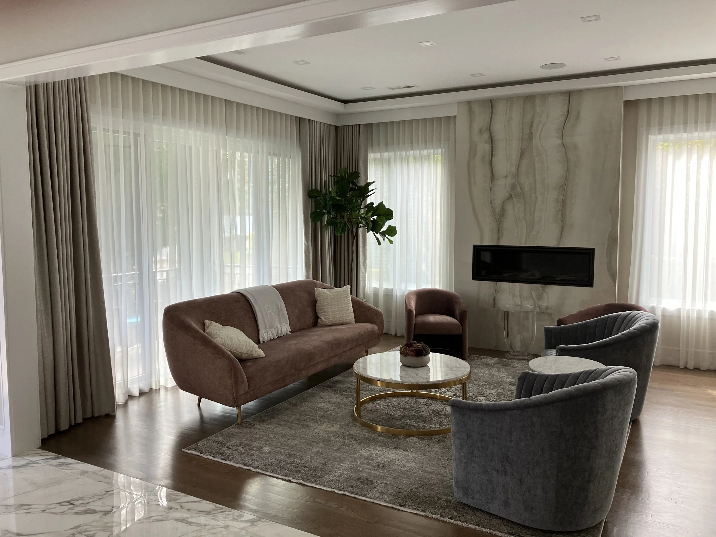 Elegant living room with mauve sofa, two grey velvet chairs, a marble coffee table with a decorative bowl, a flat-screen TV, gold accents, and large windows with sheer curtains.