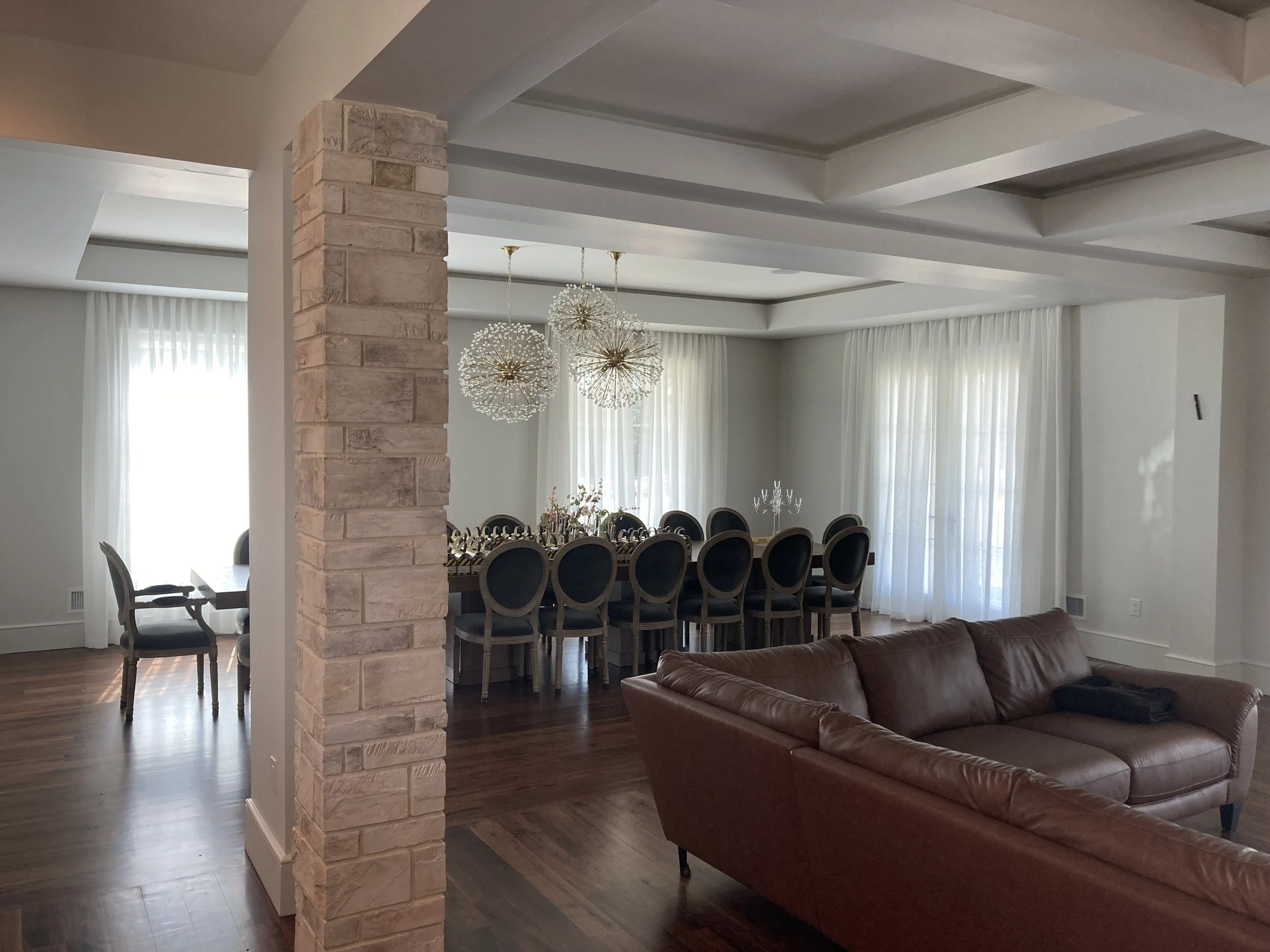 Interior of a living and dining room with hardwood floors, white walls, sheer white curtains, a brown leather couch, a dining table with black chairs, and modern chandeliers hanging from the ceiling.