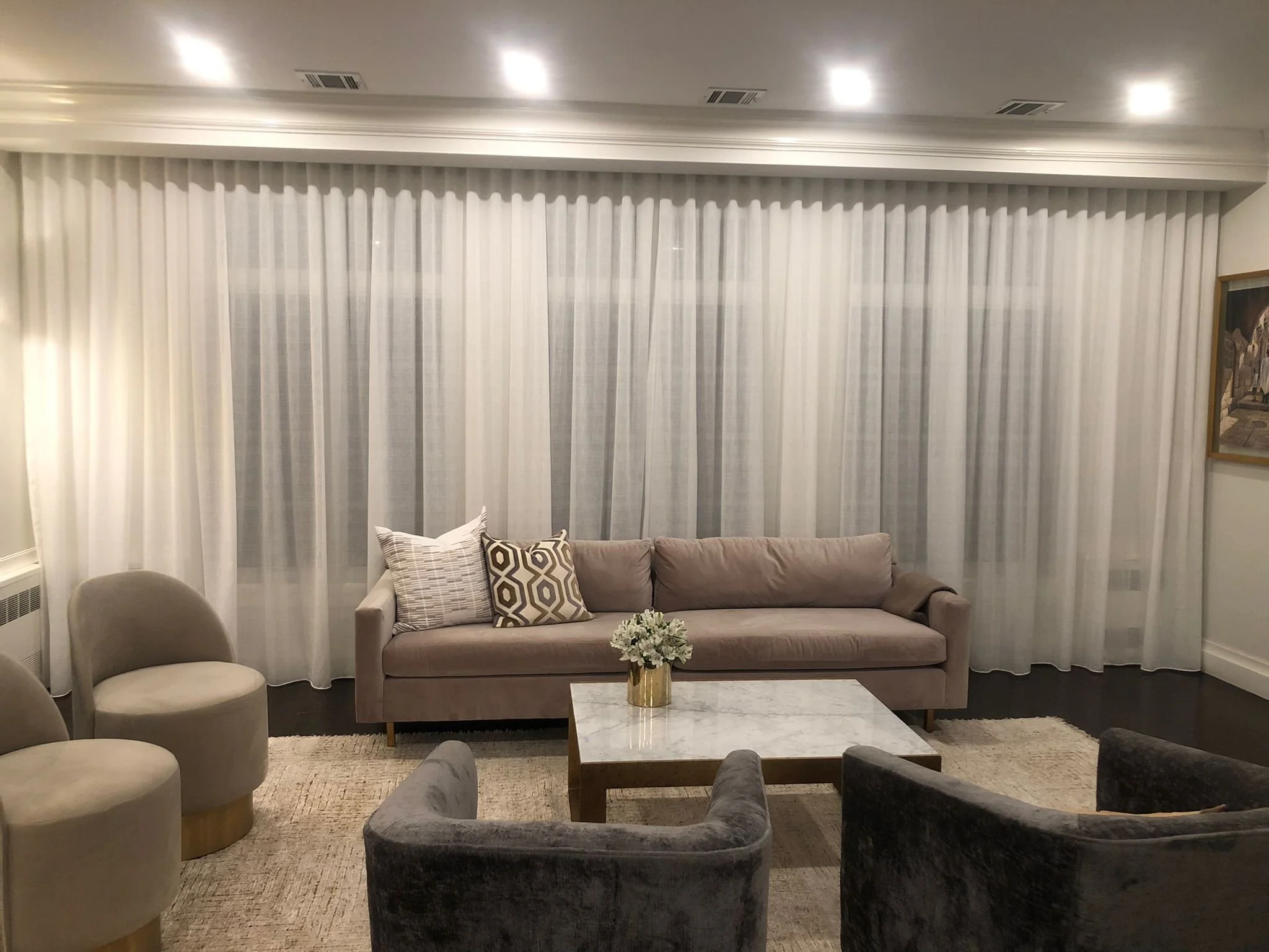Living room with light beige sofa, patterned pillows, glass coffee table with flower arrangement, wall of white curtains, and framed painting.