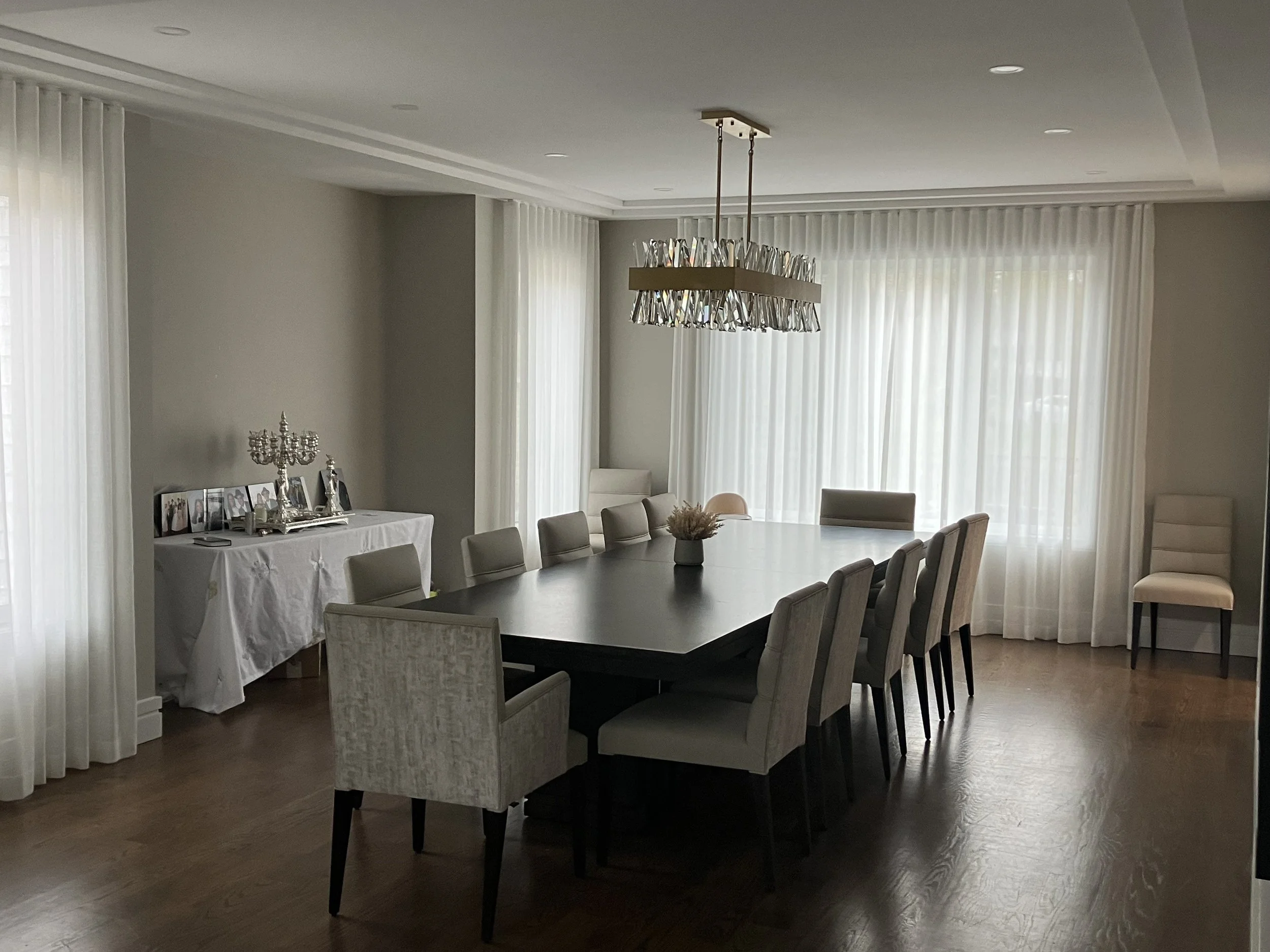 Dining room with a long dark wood table surrounded by white upholstered chairs, a chandelier hanging above, large windows with white curtains, and a side table with framed photographs and a decorative candelabrum.