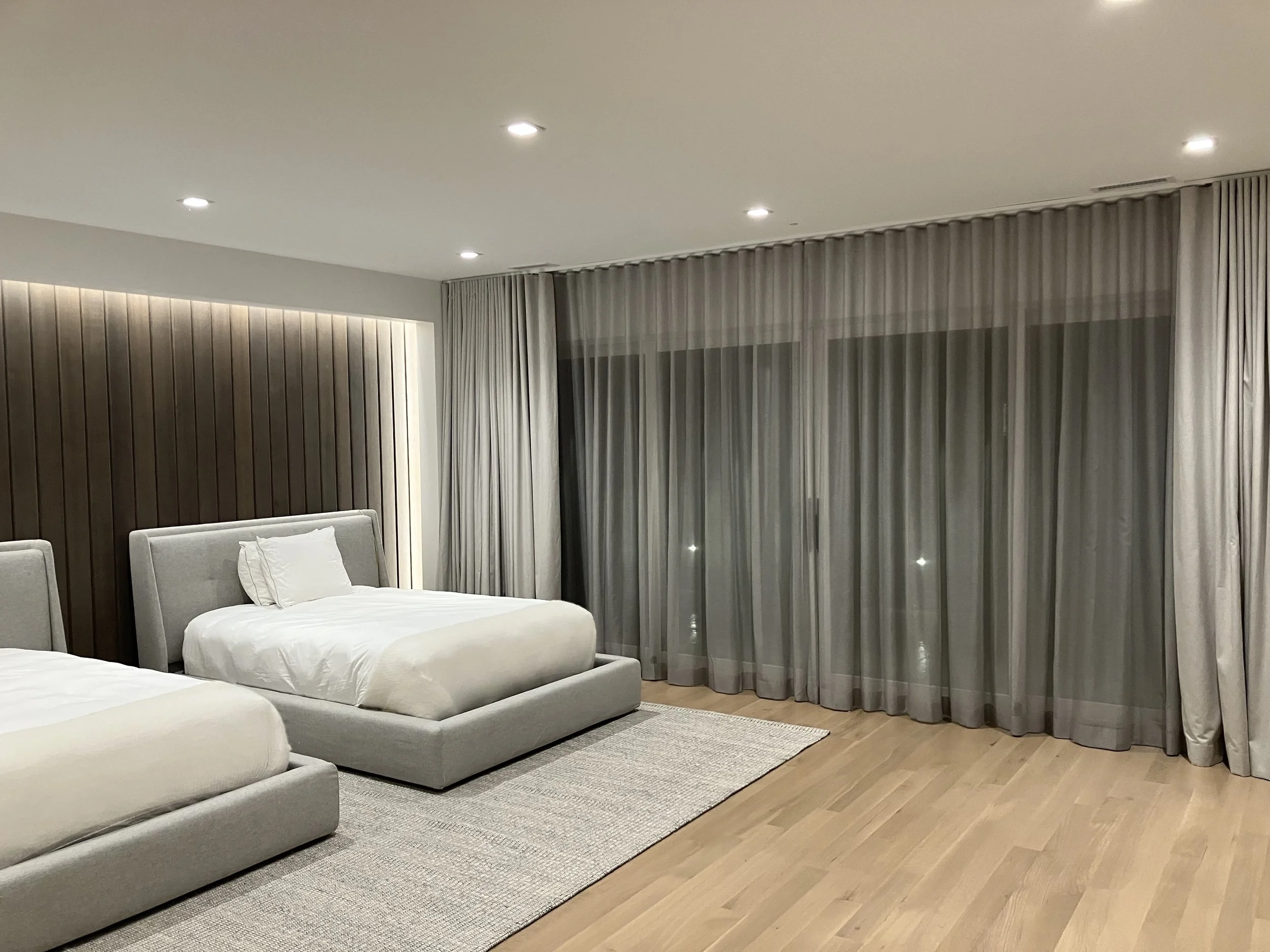 A modern hotel room with two beds, a wooden accent wall, gray curtains covering a large window, and a light-colored rug on wooden flooring.