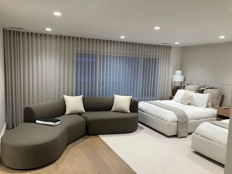 Modern bedroom with gray and beige furniture, including a curved gray sectional sofa and two beds with white bedding, situated near large window with sheer curtains.