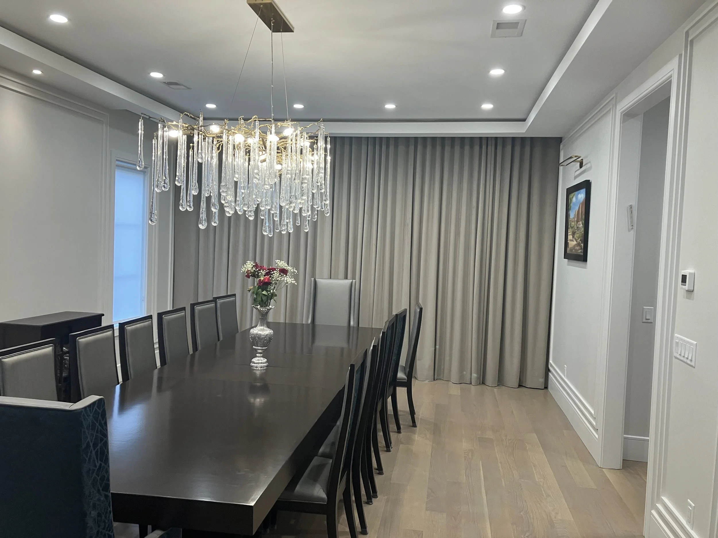 Elegant dining room with a long dark wooden table, black and silver chairs, a chandelier with hanging glass elements, a vase with flowers, beige curtains, a framed painting, and hardwood flooring.