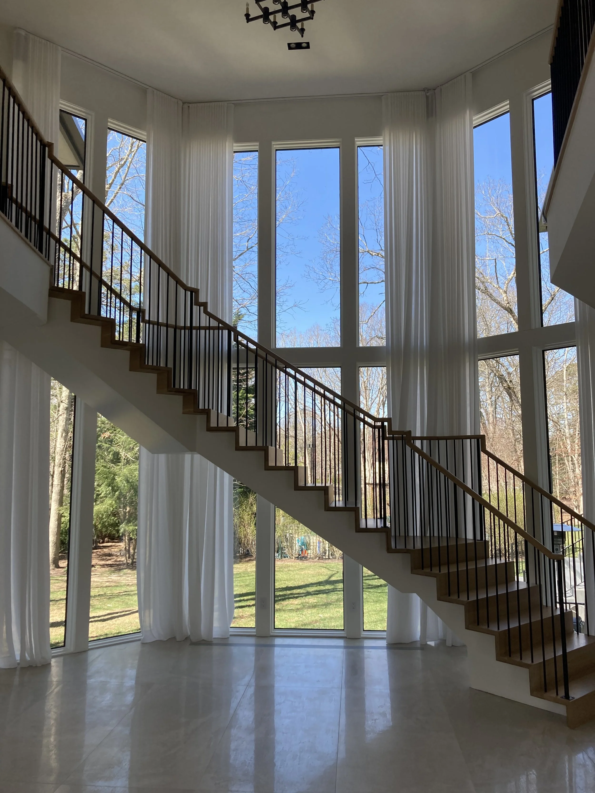 Interior view of a modern staircase inside a house, featuring a large floor-to-ceiling window with sheer white curtains, overlooking a backyard with trees, green grass, and a blue sky.