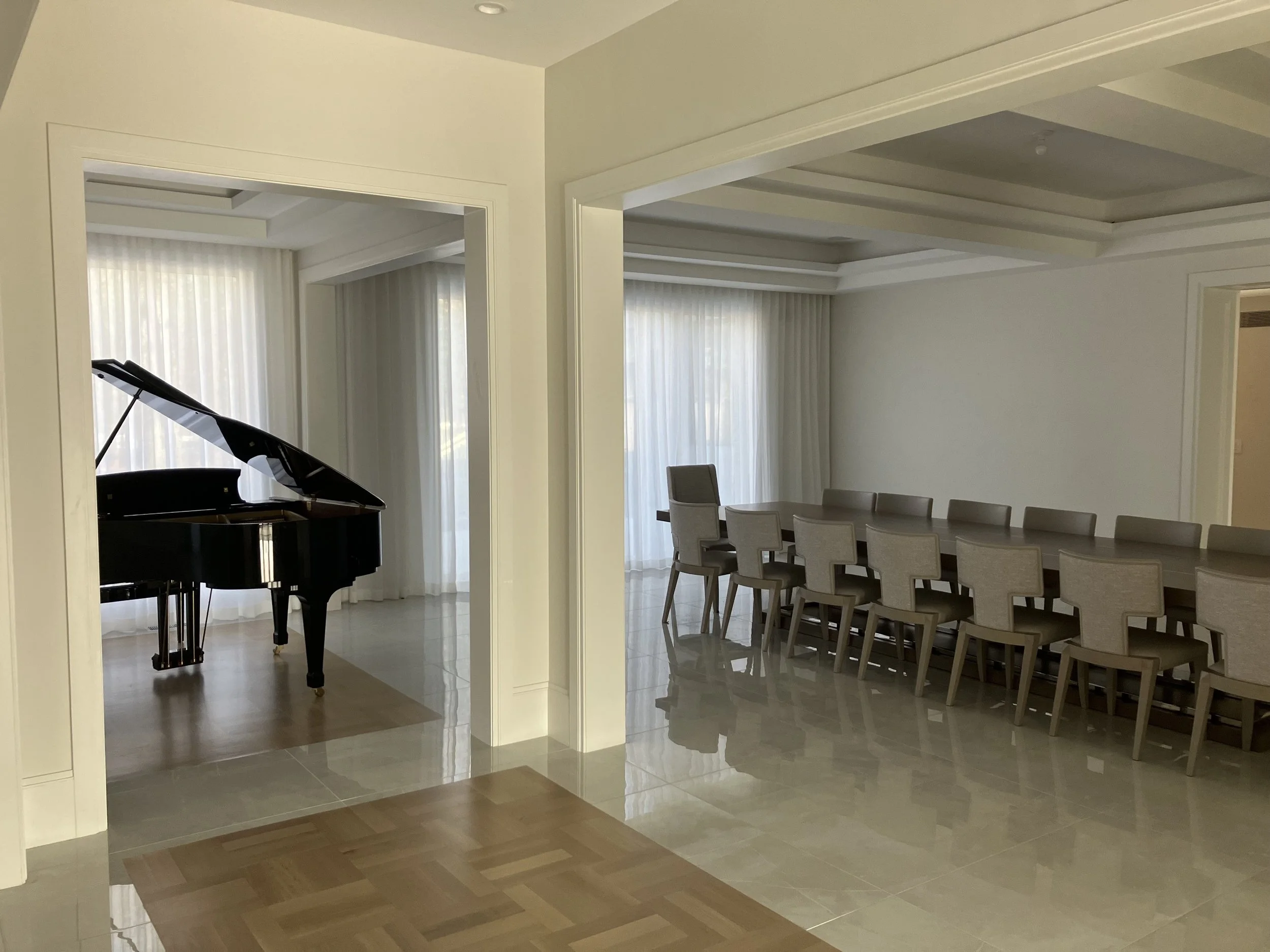 A spacious room featuring a black grand piano and a long dining table with beige chairs, with large windows covered by sheer curtains, and polished tile floors.