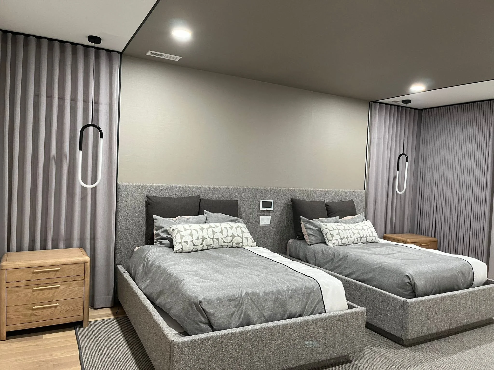 Modern hotel room with two beds, gray bedding, black and gray pillows, wooden bedside tables, gray curtains, and contemporary hanging lamps.