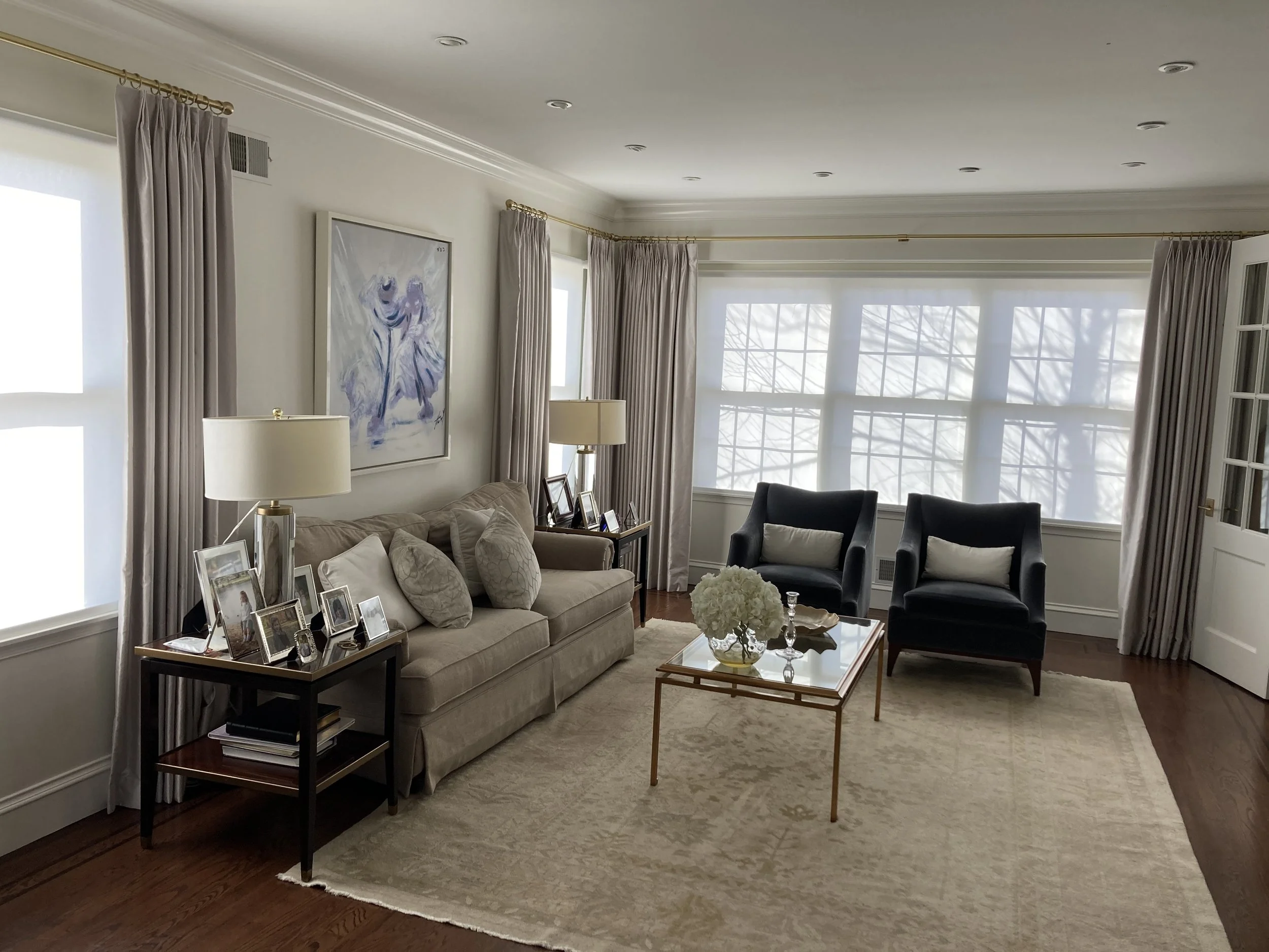 Living room with beige sofa, two black armchairs, glass coffee table with white flowers, side tables with lamps, framed photographs, artwork, and closed curtains on large windows.