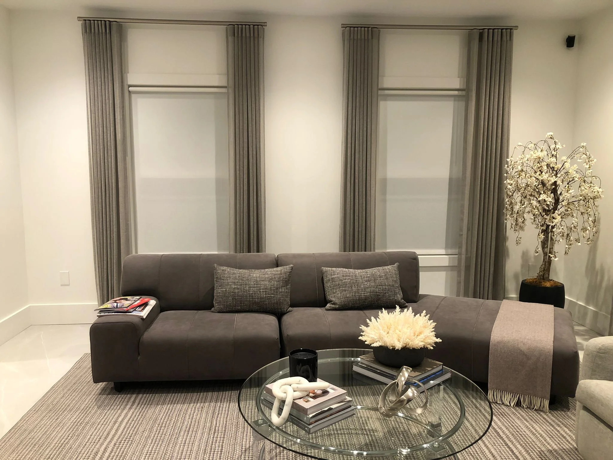 Living room with a gray sectional sofa, gray curtains, a round glass coffee table with books, decorative objects, and a flower arrangement, and a white tree-shaped plant in a black pot.