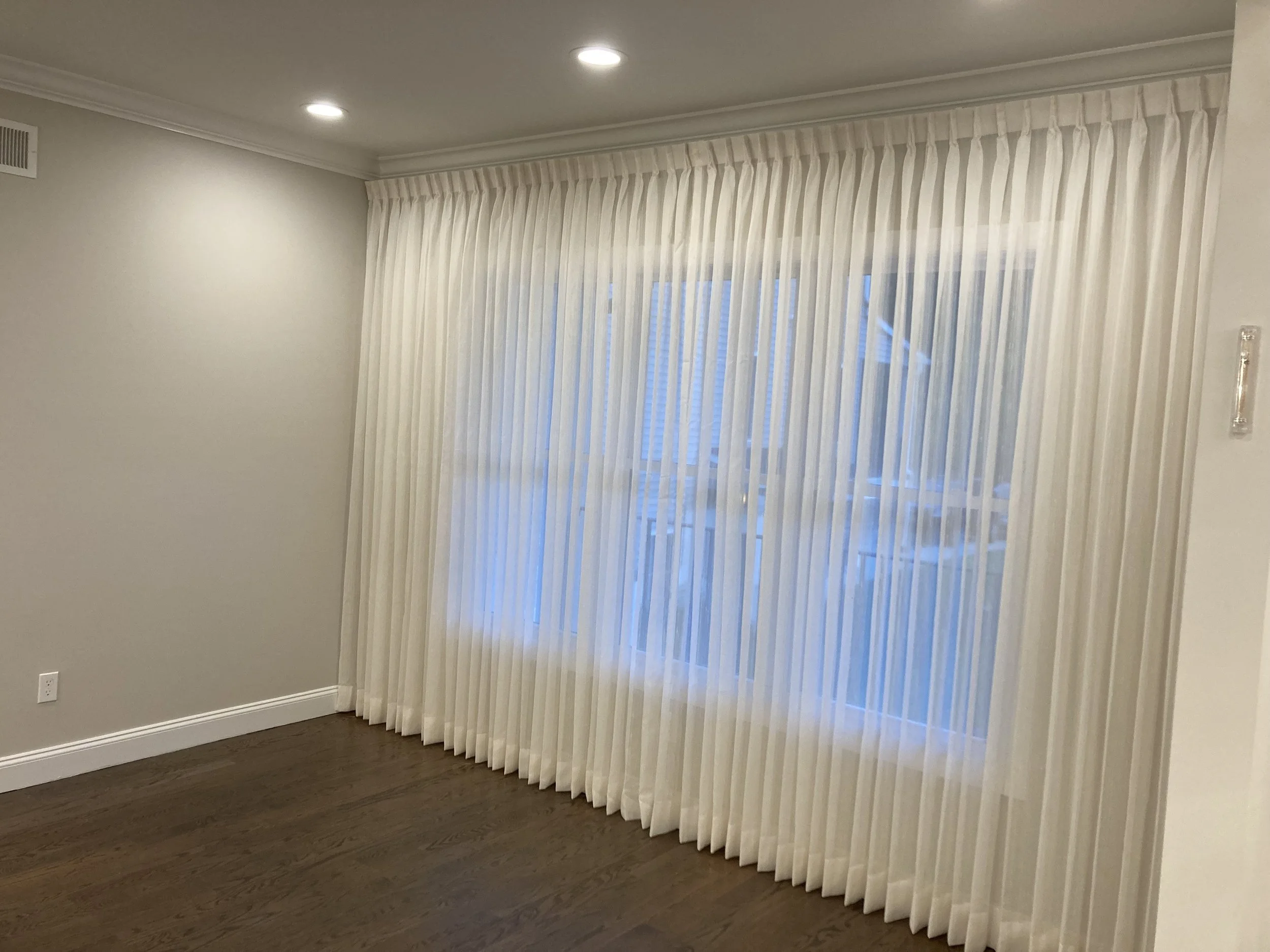 Room with white curtains covering a large window, dark hardwood floor, and recessed ceiling lights.