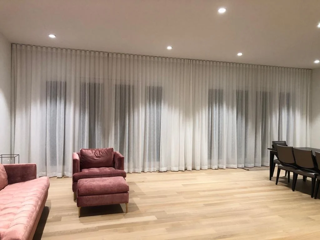 A spacious room with smoke-colored curtains covering large windows, a pink tufted sofa, a matching pink armchair with an ottoman, and a dining table with six black chairs. Recessed ceiling lights illuminate the room. The floor is light wood.